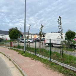 Usługi Brukarsko-Ogrodzeniowe UZBUD Marcin Guz - Nowo zamontowane, zielone ogrodzenie panelowe z podmurówką betonową, oddzielające chodnik z czerwonej kostki od terenu zielonego z młodymi drzewkami i zaparkowanymi autami.