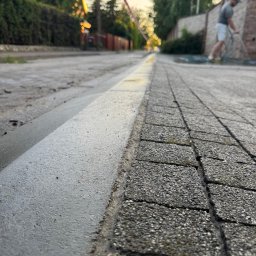 Usługi Brukarsko-Ogrodzeniowe UZBUD Marcin Guz - Podniesiony wjazd z kostki brukowej, widok z poziomu gruntu. W tle mężczyzna myje nawierzchnię, a w oddali widoczne domy i zieleń.