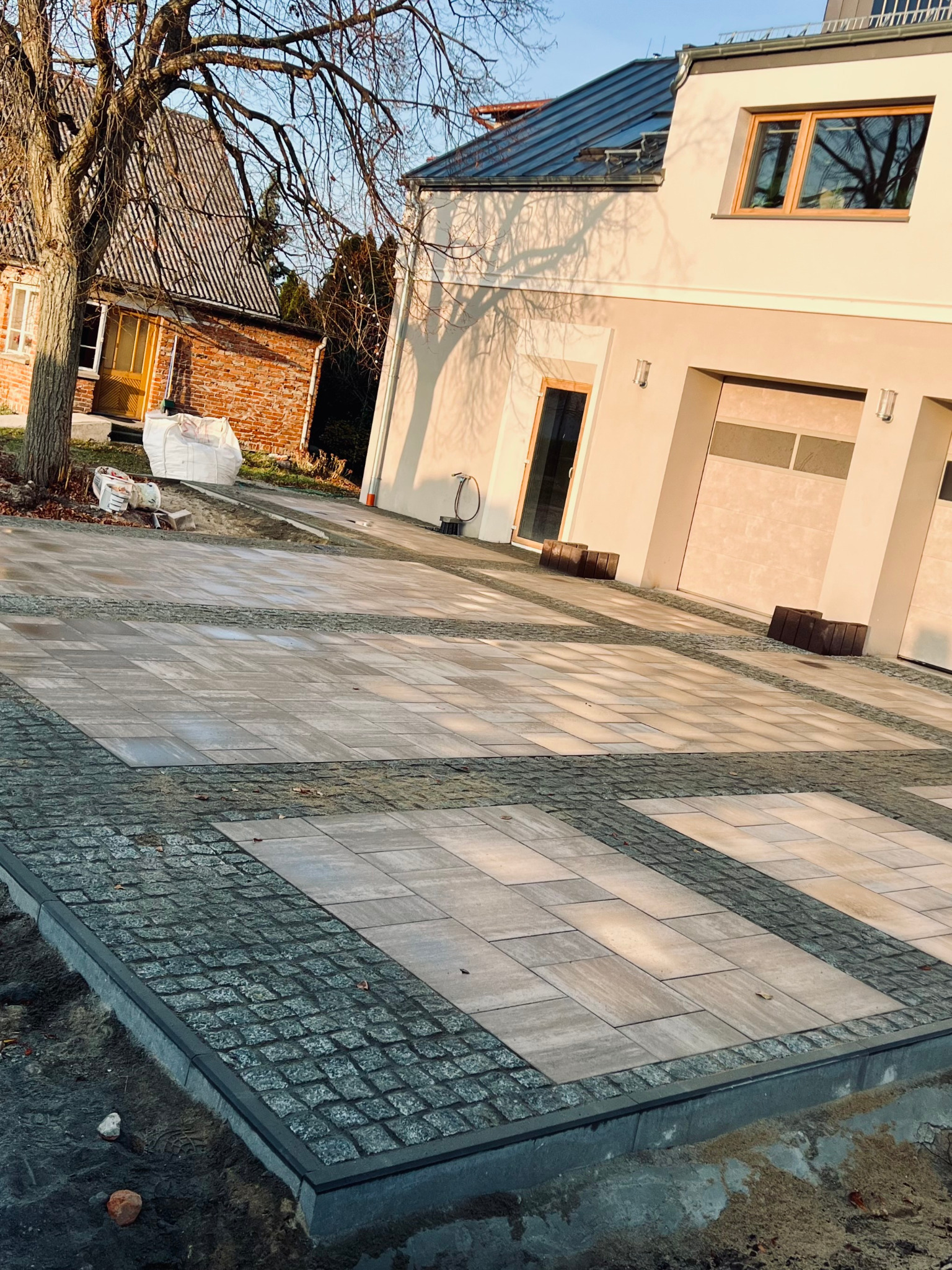 Ułożona kostka brukowa Toledo Polbruk i granit cięto-łupany 8x8, 10x10 na podjeździe. Widok na dom z jasną elewacją i garażem. Częściowo widoczne drzewo.
