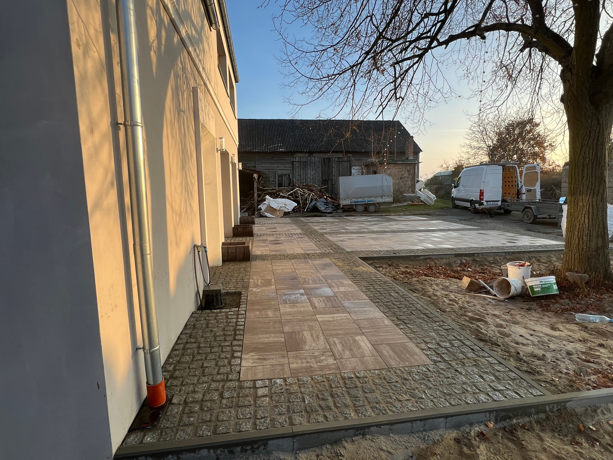 Ułożona kostka brukowa Toledo Polbruk z obrzeżem z kostki granitowej cięto-łupanej 8x8/10x10 na posesji z budynkiem gospodarczym w tle, w jesiennej scenerii.