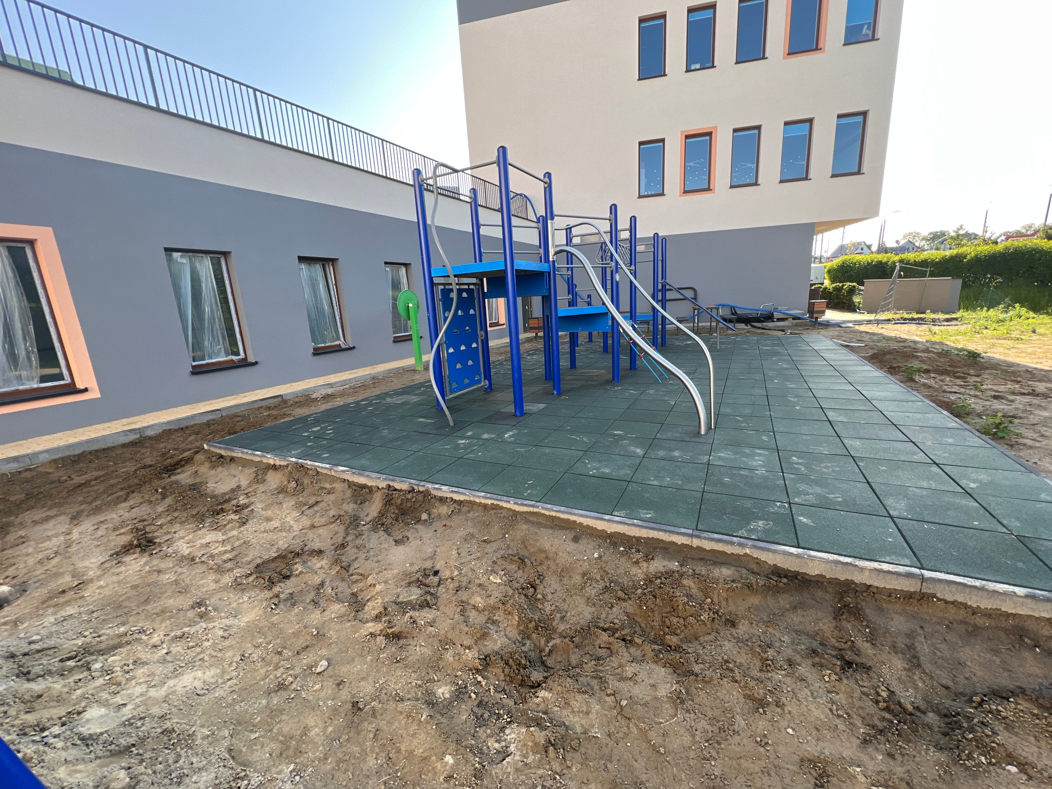 Nierówny teren przed placem zabaw z niebieskimi elementami i zieloną nawierzchnią. Budynek w tle z szarymi i beżowymi ścianami oraz oknami z brązowymi ramami.