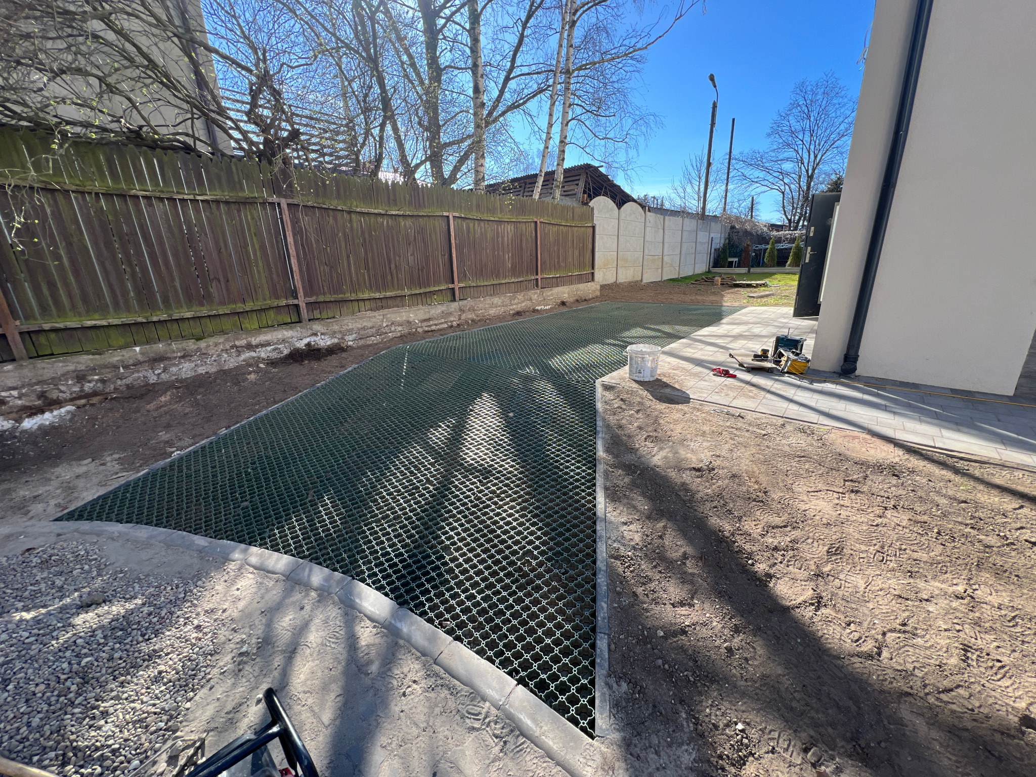 Przygotowany teren pod kostkę z siatką wzmacniającą, widoczne narzędzia i wiadro, w tle ogrodzenie drewniane i betonowe płyty. Słoneczny dzień.