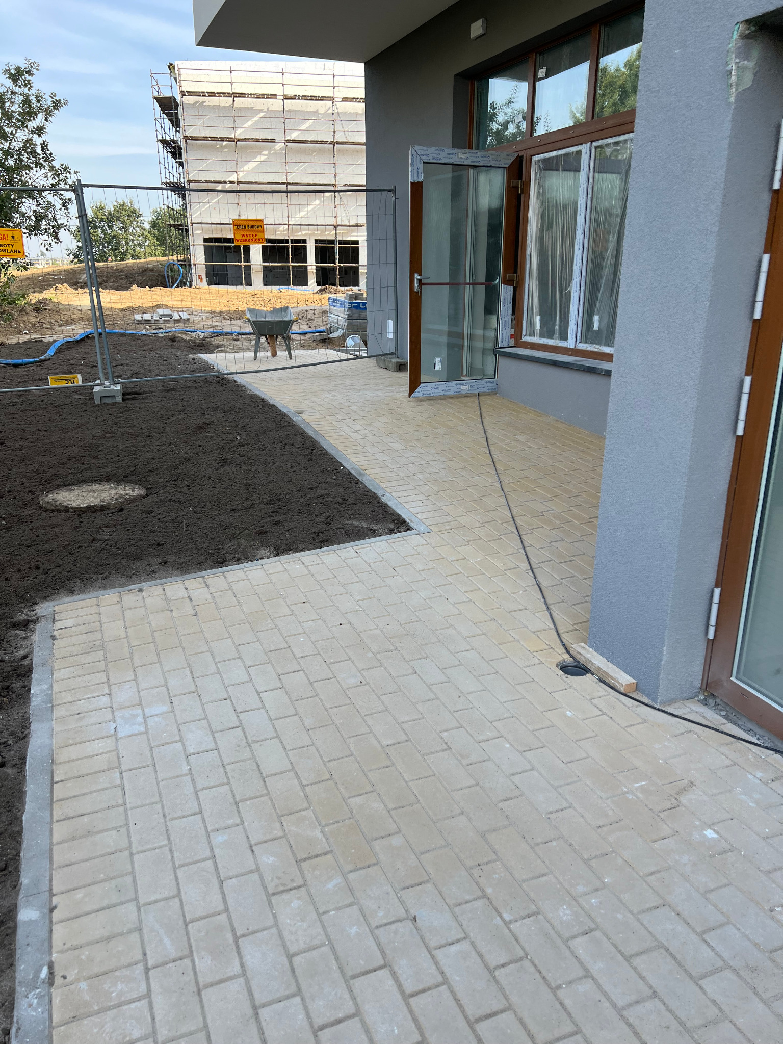 Jasna kostka brukowa tworzy chodnik przy budynku z otwartymi drzwiami. W tle plac budowy z rusztowaniami i przygotowany teren pod trawnik.
