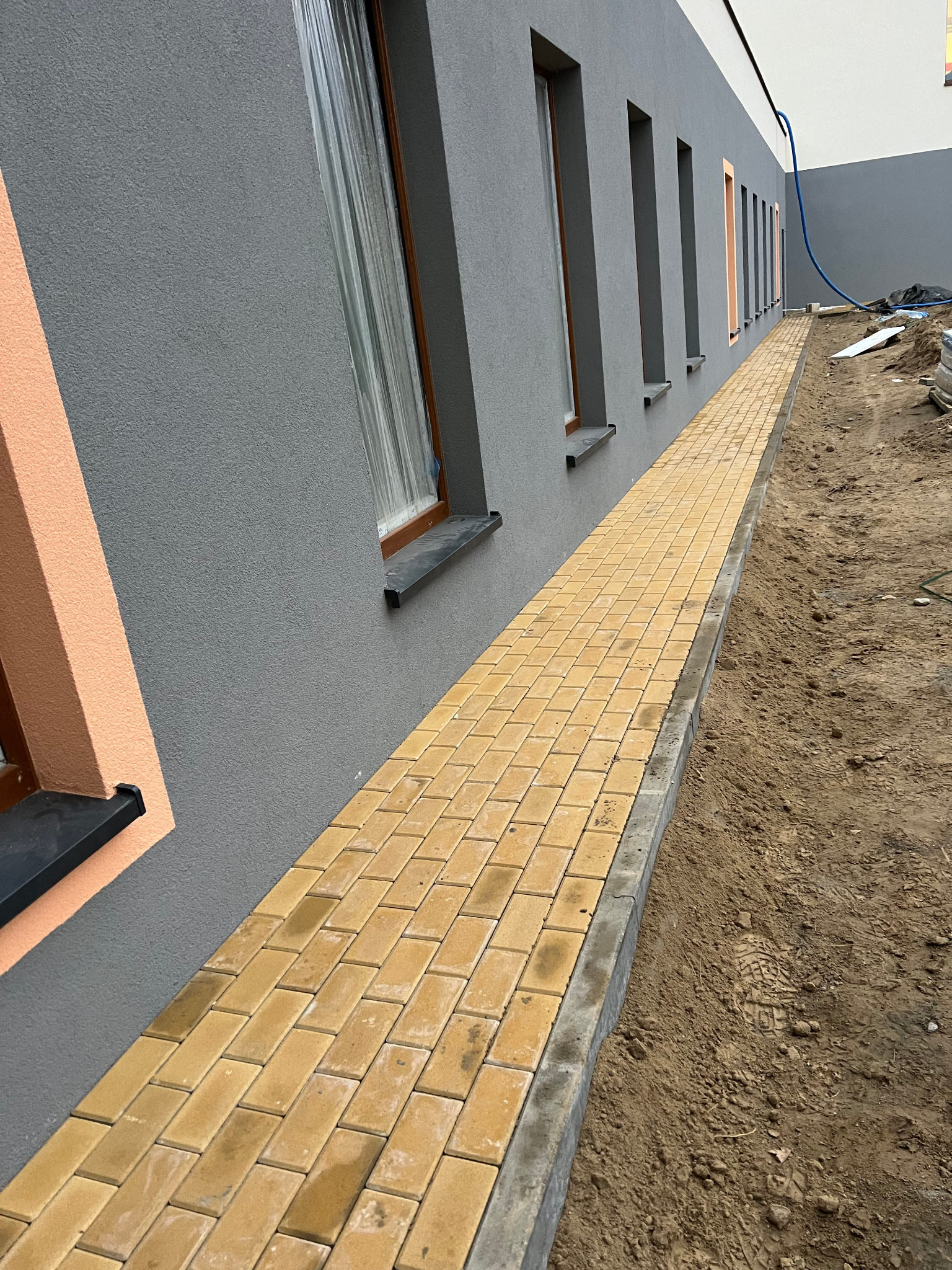 Żółta kostka brukowa tworzy chodnik wzdłuż szarej ściany budynku z oknami. Widoczny fragment terenu przed budynkiem w trakcie prac wykończeniowych.