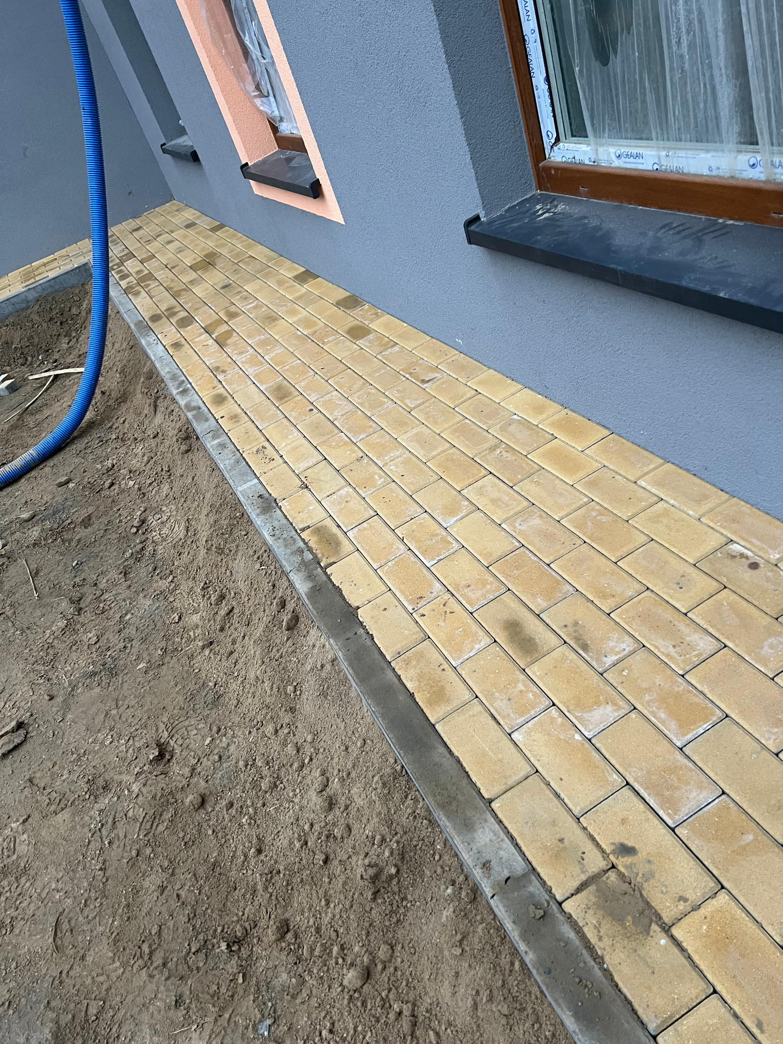 Żółta kostka brukowa ułożona wzdłuż ściany budynku z oknami. Widoczny fragment niezagospodarowanego terenu i niebieski wąż. Praca w toku, etap układania chodnika.