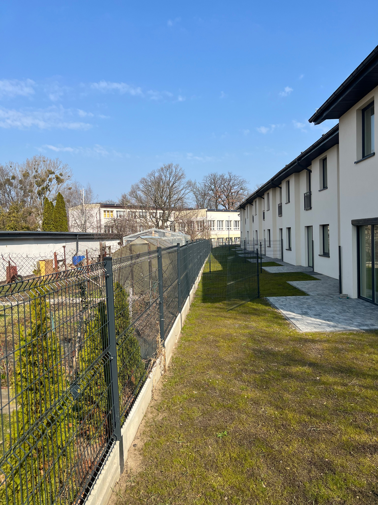 Nowoczesne ogrodzenie panelowe w kolorze grafitowym, oddzielające posesje szeregowych domów z kostką brukową i trawnikiem. Błękitne niebo w tle.