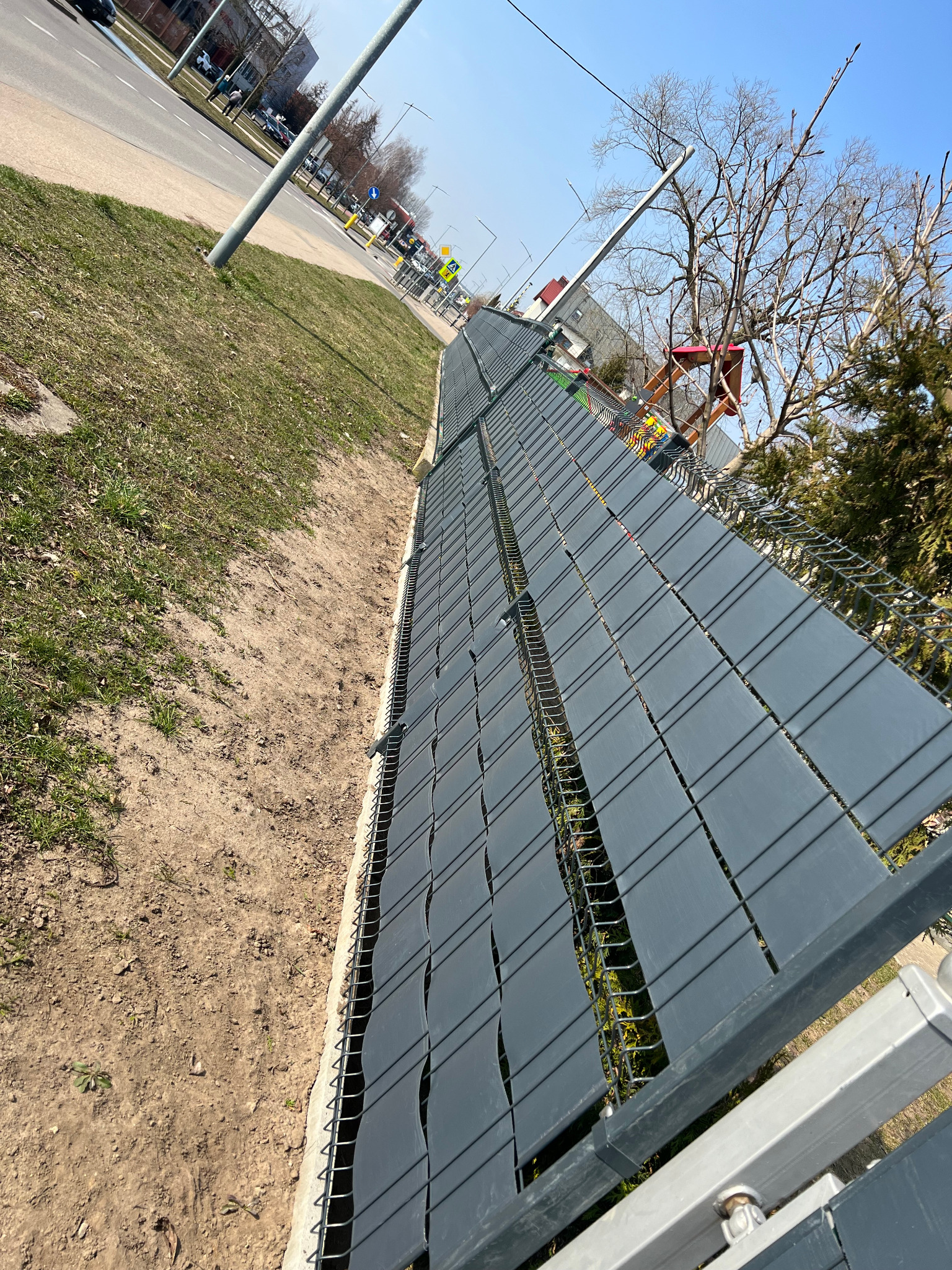 Długi, szary panelowy płot z taśmą ogrodzeniową, widziany z perspektywy bocznej, oddzielający trawnik od ulicy w słoneczny dzień. Widoczne elementy montażowe.