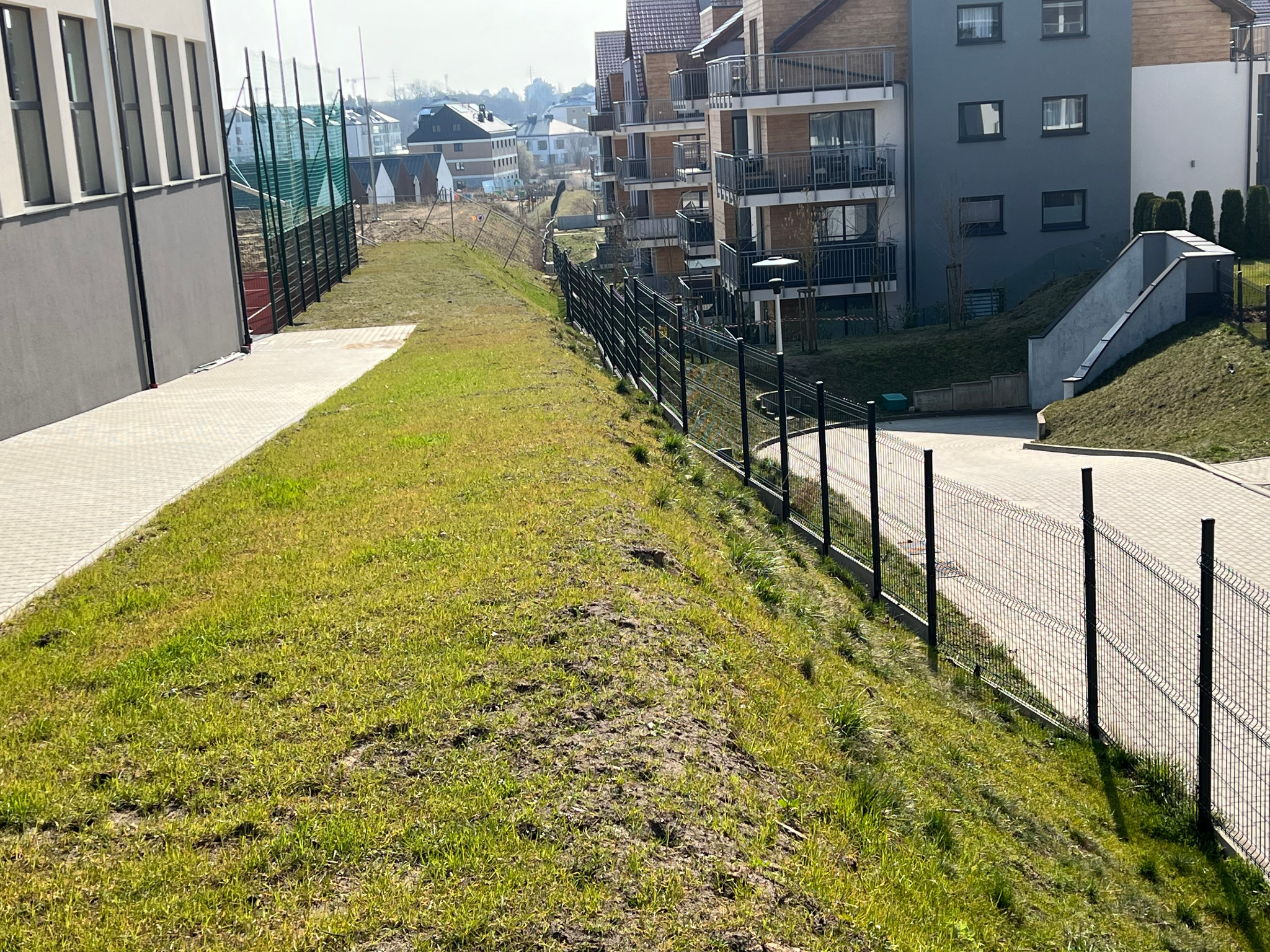 Długi, czarny panelowy płot okalający zielony teren szkoły, z budynkami mieszkalnymi w tle. Widok z perspektywy.