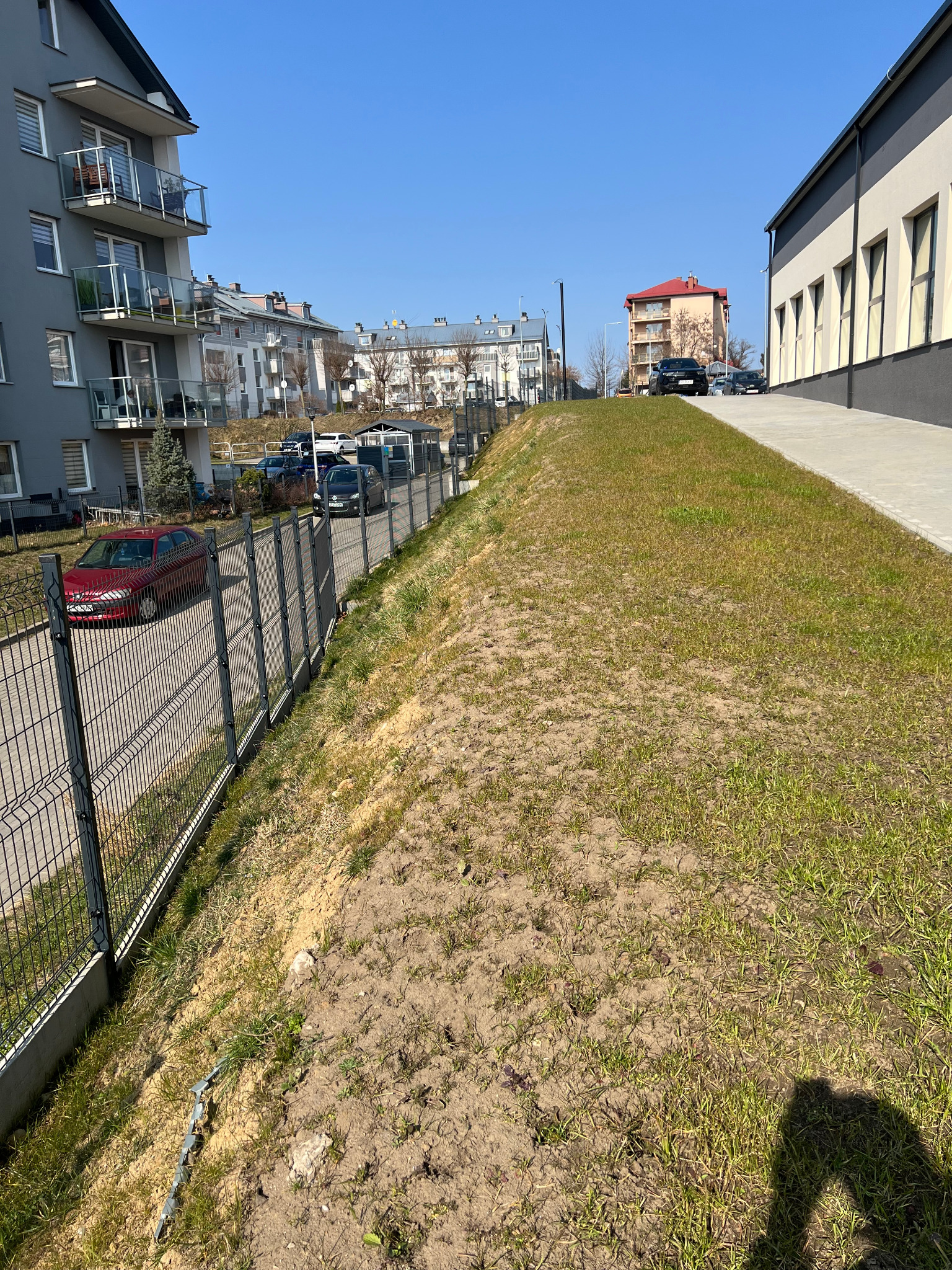 Nowoczesne ogrodzenie panelowe z metalu, oddzielające chodnik od terenu zielonego. W tle budynki mieszkalne i szkoła w słoneczny dzień.