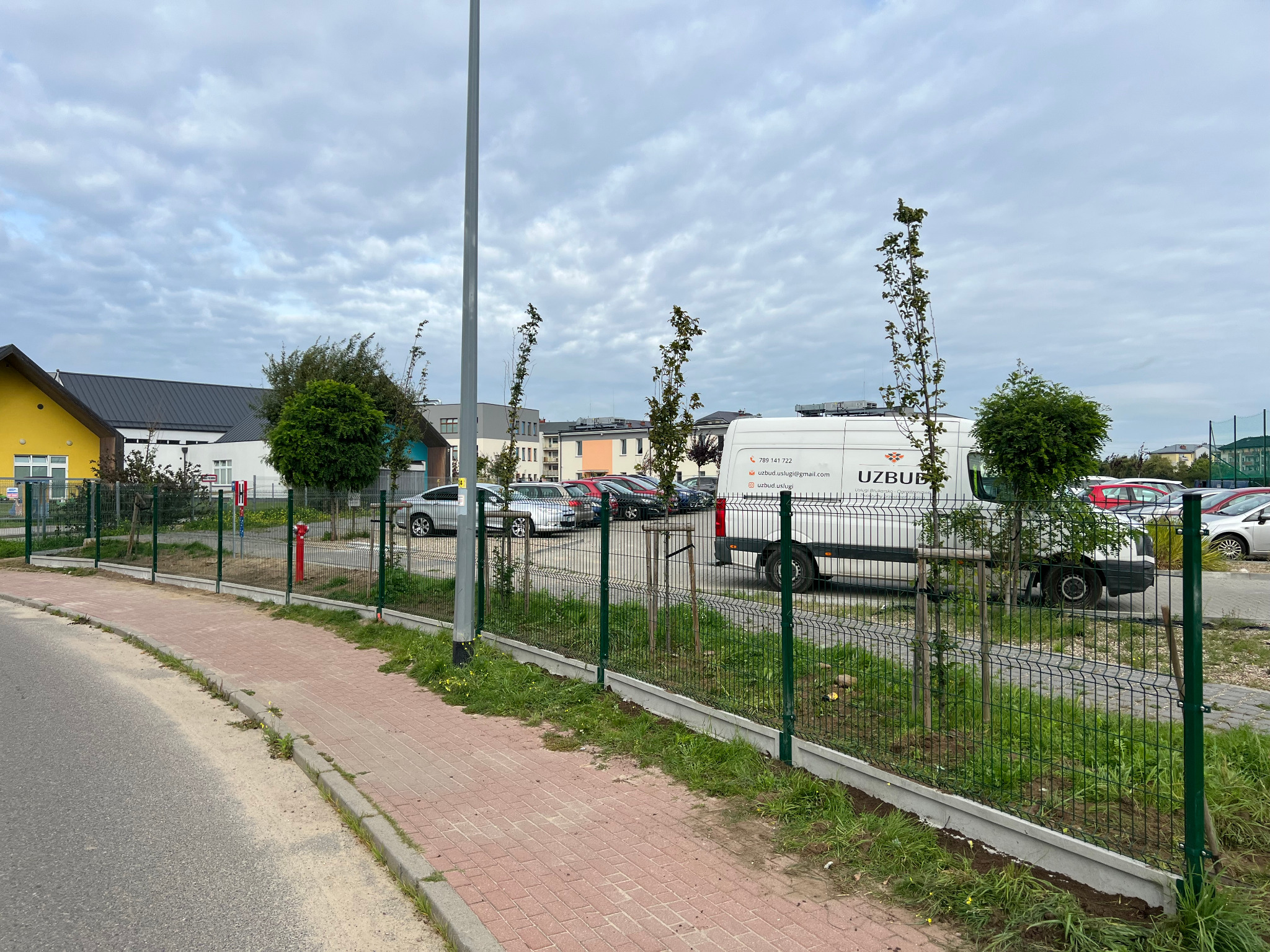 Nowo zamontowane, zielone ogrodzenie panelowe z podmurówką betonową, oddzielające chodnik z czerwonej kostki od terenu zielonego z młodymi drzewkami i zaparkowanymi autami.