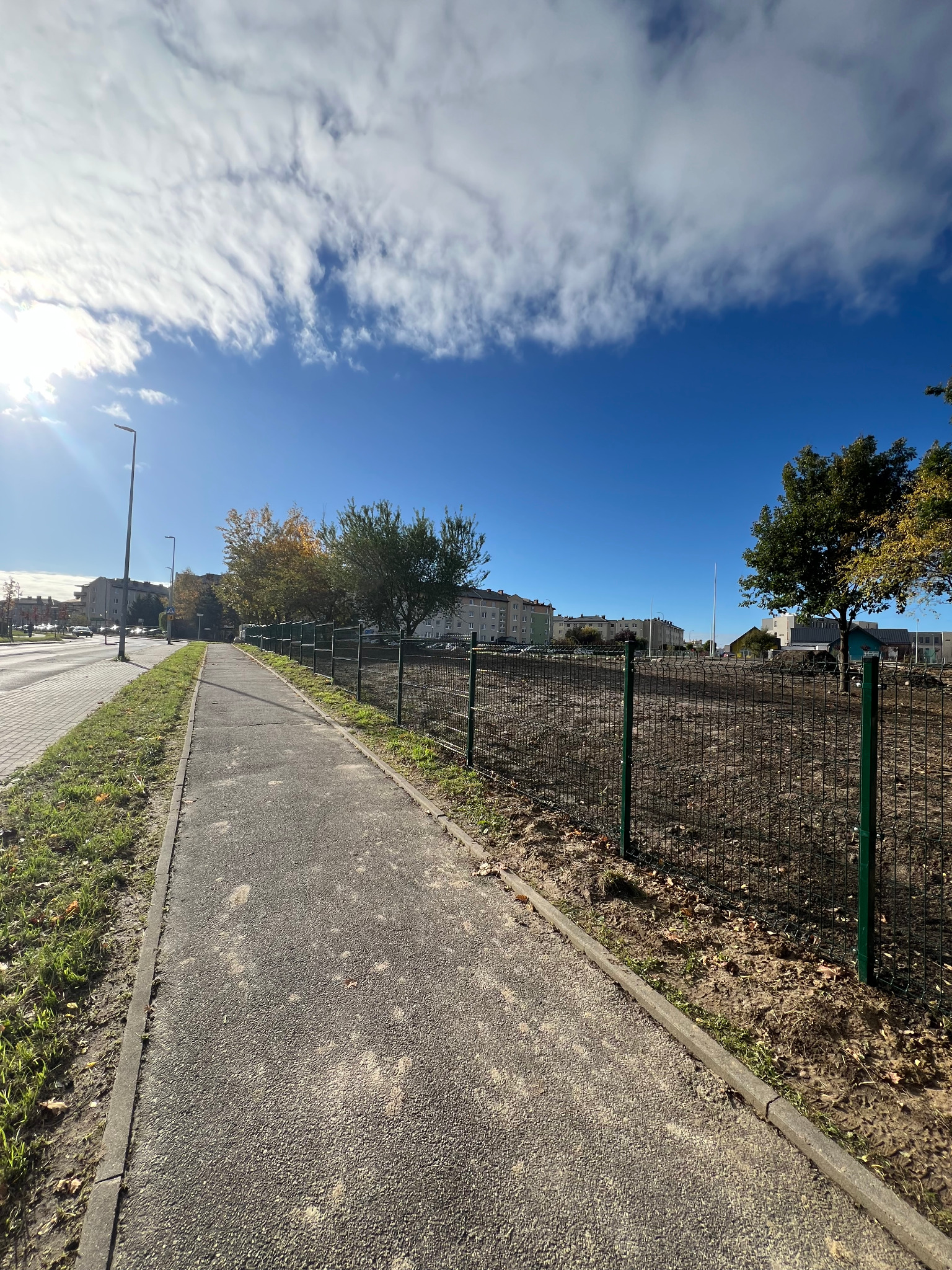 Zielone ogrodzenie z siatki otacza teren. Widok z chodnika w słoneczny dzień. W tle budynki mieszkalne i błękitne niebo z chmurami.