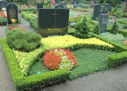 Uporządkowany grób z niskim żywopłotem, kolorowymi kwiatami i krzewami, w tle nagrobki na cmentarzu.