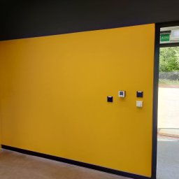 BGRM Grzegorz Chołda - Ściana pomalowana na żółto z czarnymi akcentami u góry i dołu, z zamontowanymi przełącznikami i termostatem, widok na zewnątrz przez otwarte drzwi.