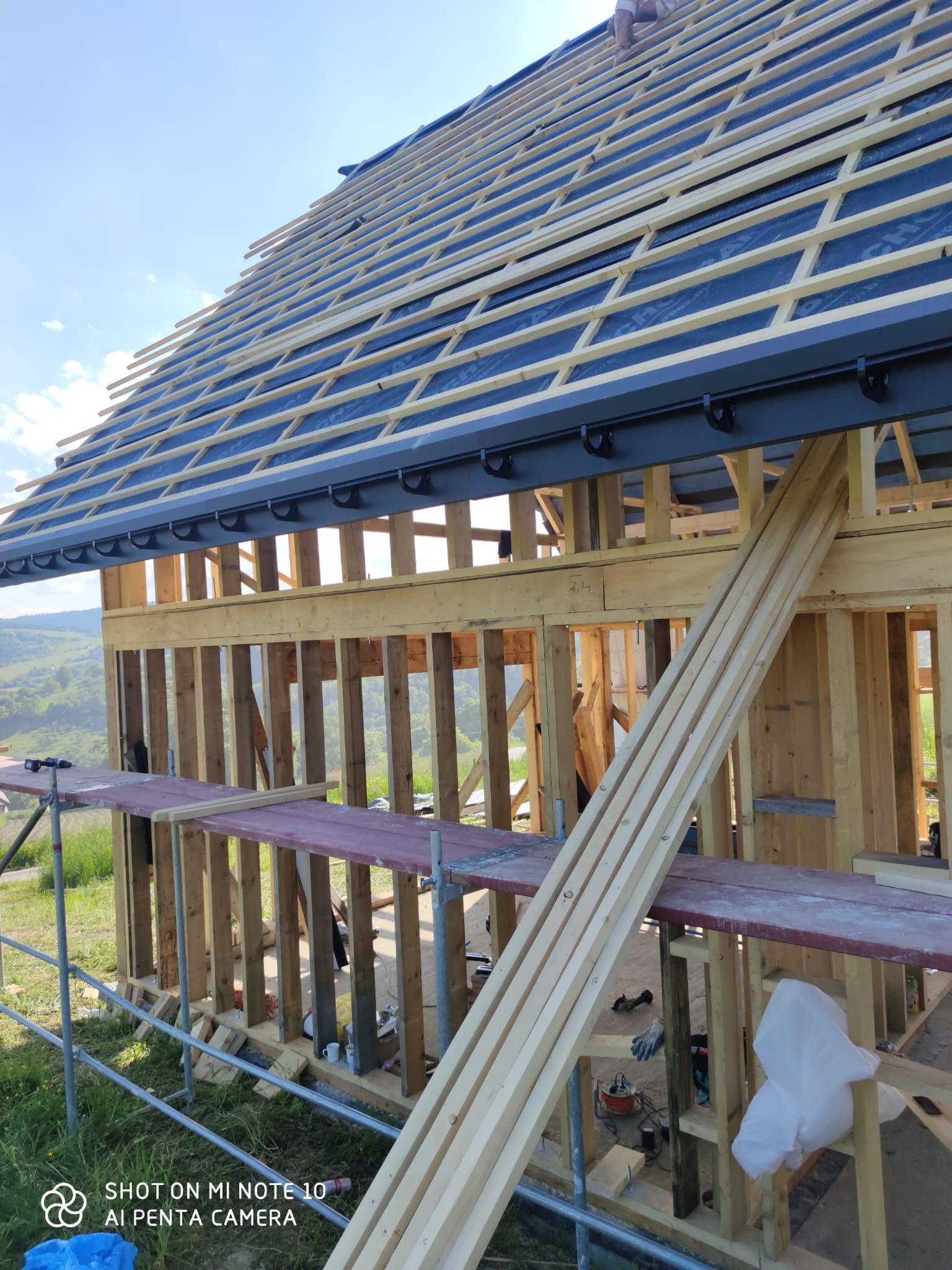 Konstrukcja drewniana domu w trakcie budowy z widoczną więźbą dachową, deskowaniem i elementami orynnowania, na tle zielonych wzgórz.