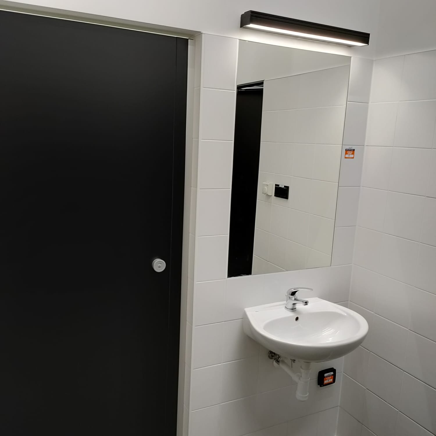Nowoczesna łazienka z czarnymi drzwiami i białą umywalką na tle białych płytek, oświetlona podłużną lampą nad lustrem.