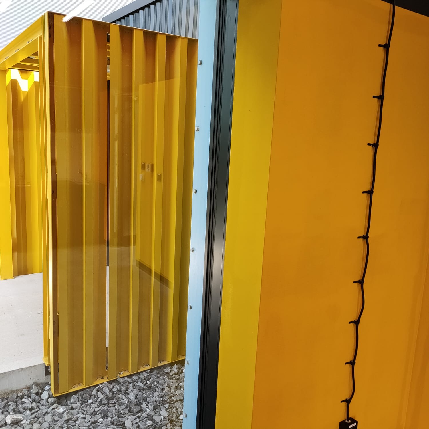 Żółta, lamelowa konstrukcja z surowymi, szarymi kamieniami u podstawy, sąsiadująca z pomarańczową ścianą z kablem elektrycznym