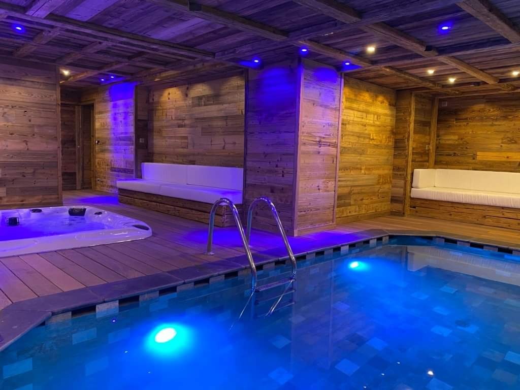 Kryty basen i jacuzzi w pomieszczeniu wykończonym drewnem, z niebieskim oświetleniem punktowym na suficie i pod wodą, tworzącym relaksującą atmosferę.