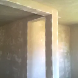 Świeżo nałożony, jasny tynk maszynowy na ścianach wewnętrznych i futrynie drzwiowej w trakcie schnięcia, widoczne nierówności i jaśniejsze plamy, sufit również pokryty tynkiem.