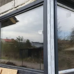 Nowe okno narożne z zamontowaną zewnętrzną roletą w szarej obudowie, widok na ceglany mur w trakcie budowy.