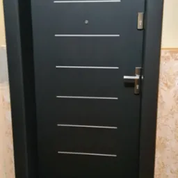 Ciemne, nowoczesne drzwi wejściowe z poziomymi, jasnymi akcentami, zamontowane w odnawianym wnętrzu z widocznymi śladami prac remontowych na ścianie.