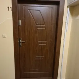 Ciemnobrązowe drzwi wejściowe do mieszkania z numerem 11 na ścianie, widoczne z bliska na tle beżowej klatki schodowej, z fragmentem drabiny malarskiej w prawym dolnym rogu.