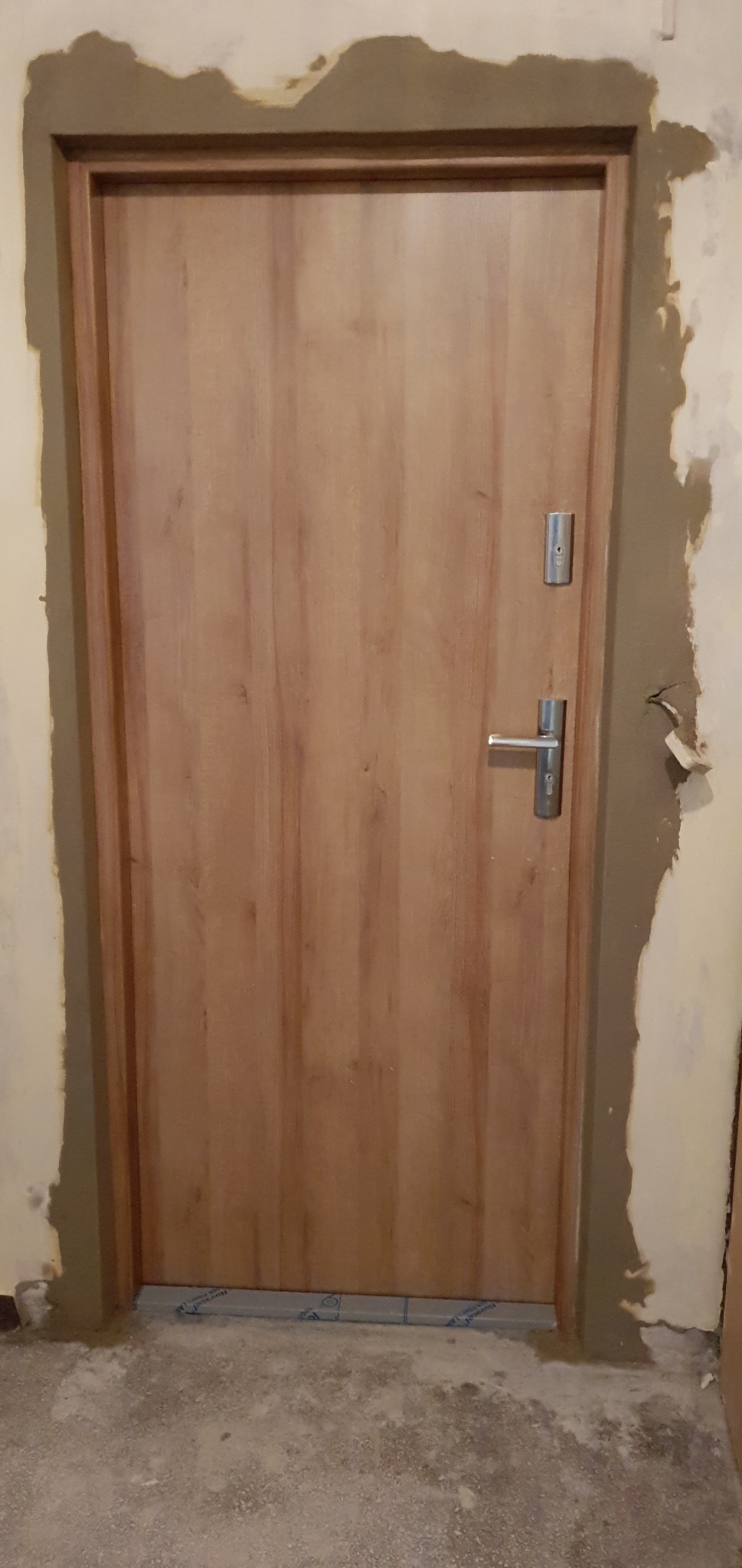Drewnopodobne drzwi wejściowe z zamkiem i klamką, osadzone w ościeżnicy z widocznymi śladami zaprawy murarskiej na ścianie i folią ochronną na progu.