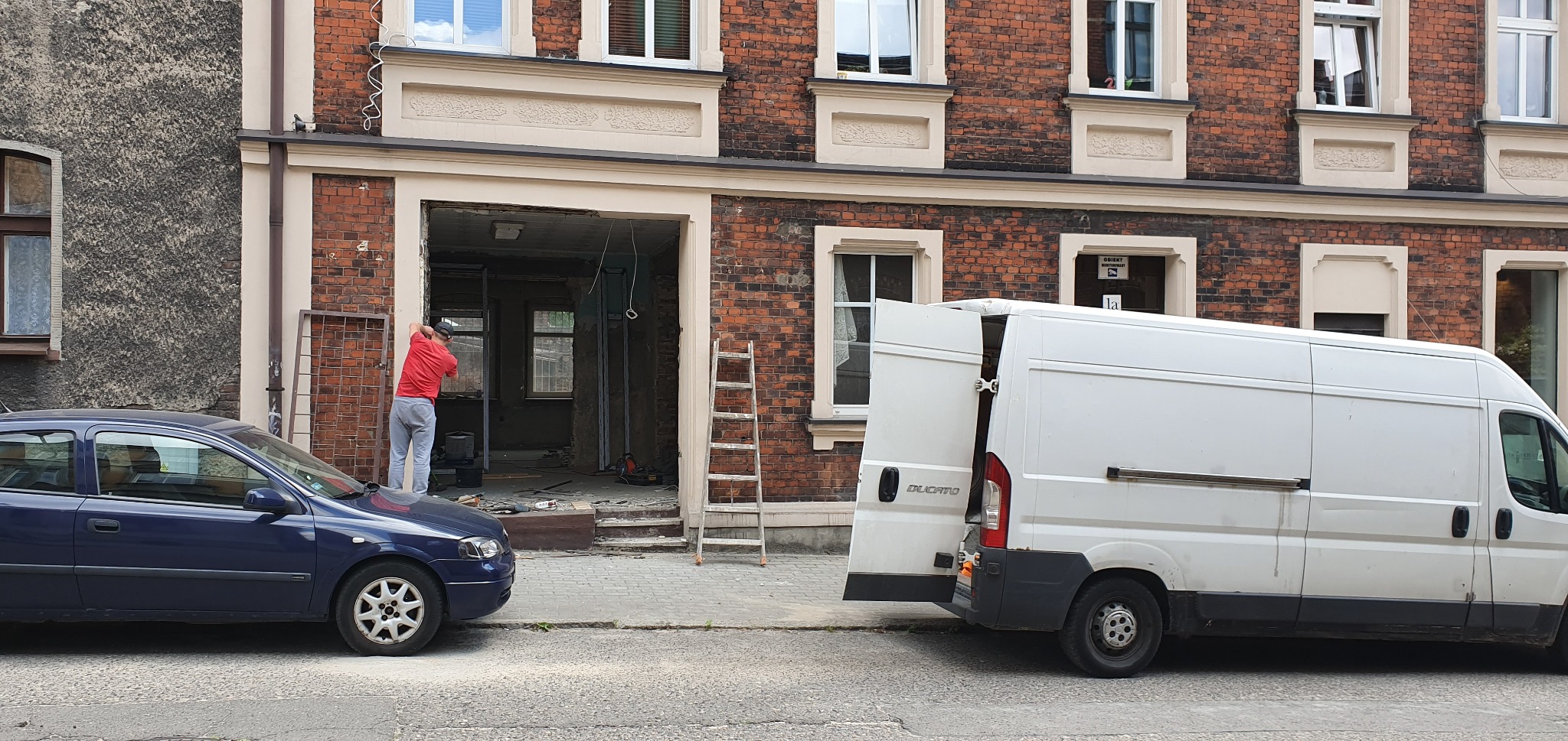 Pracownik w czerwonej koszulce montuje metalową ramę w otworze wejściowym budynku z cegły, obok zaparkowany niebieski samochód i biały van z otwartymi drzwiami.