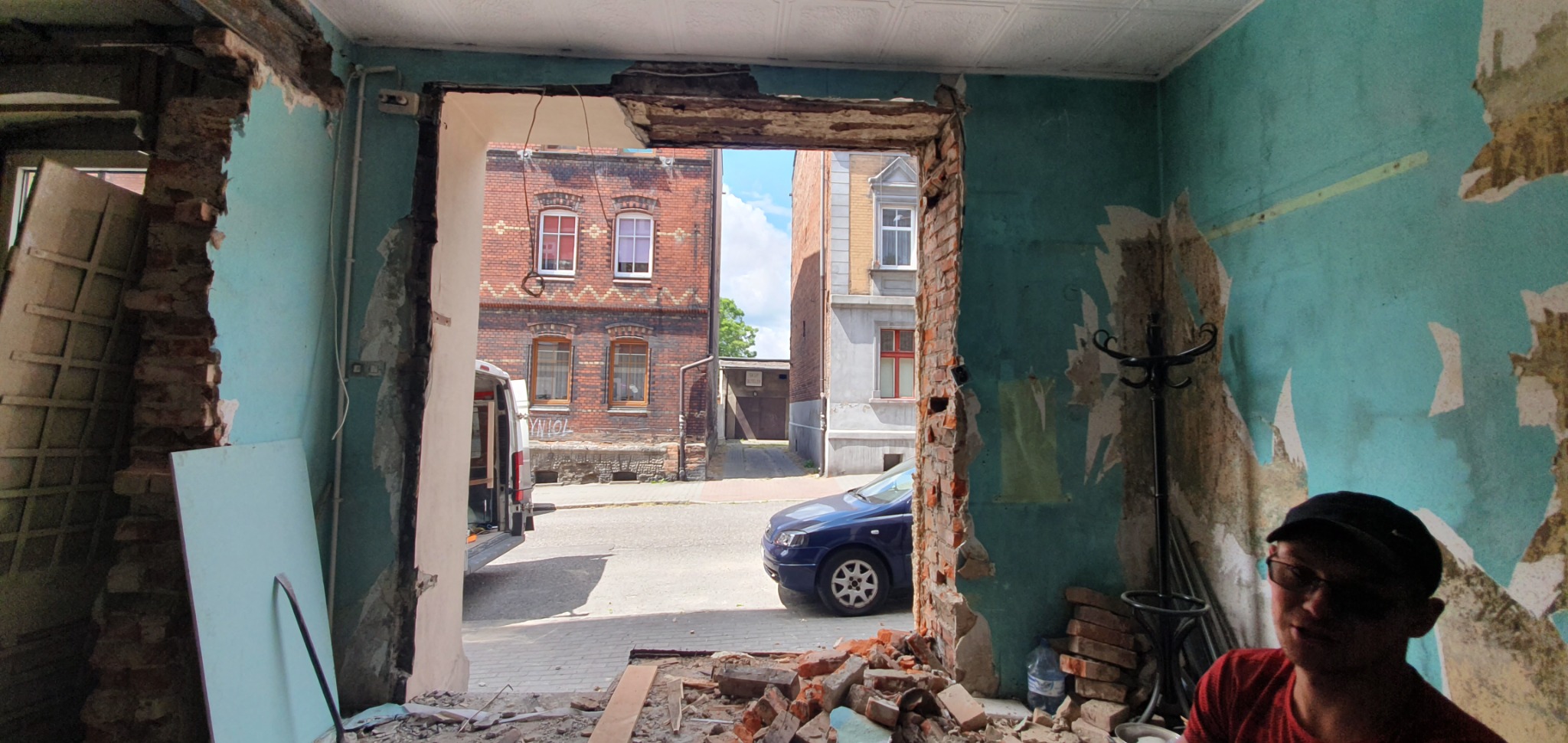 Wnętrze w trakcie remontu: zdemontowane fragmenty ściany odsłaniają cegły i widok na ulicę z zaparkowanym niebieskim samochodem oraz budynkami w tle.