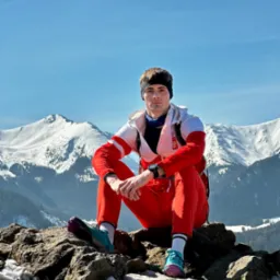 Młody mężczyzna w czerwonym dresie i sportowych butach siedzi na skalistym szczycie góry z widokiem na ośnieżone szczyty w tle.