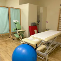 Jasne wnętrze gabinetu z profesjonalnym stołem do zabiegów, krzesłem do masażu, dużą niebieską piłką do ćwiczeń i drabinką gimnastyczną.