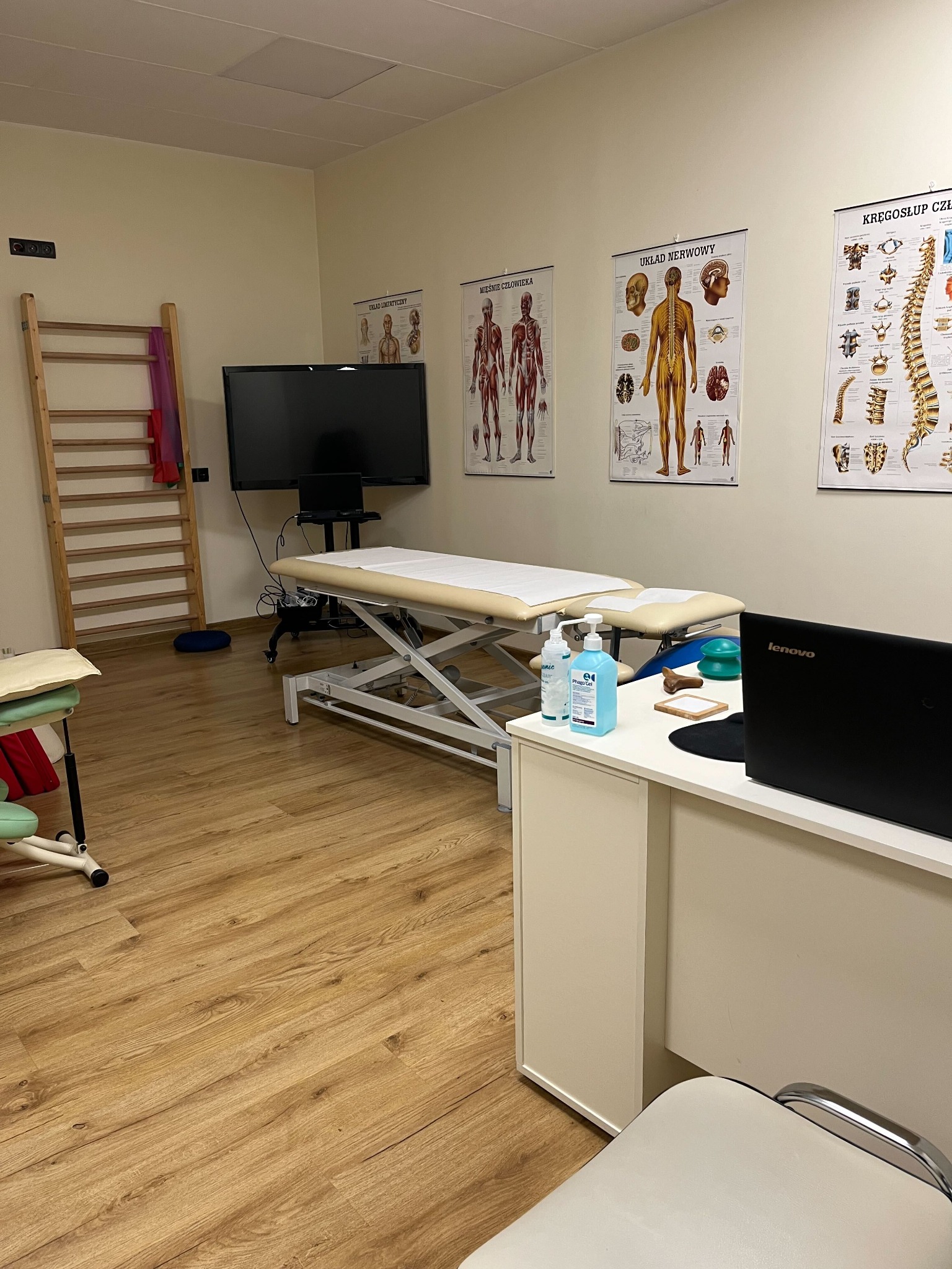 Gabinet z kozetką, drabinką rehabilitacyjną, biurkiem z laptopem Lenovo i plakatami anatomicznymi na ścianie.