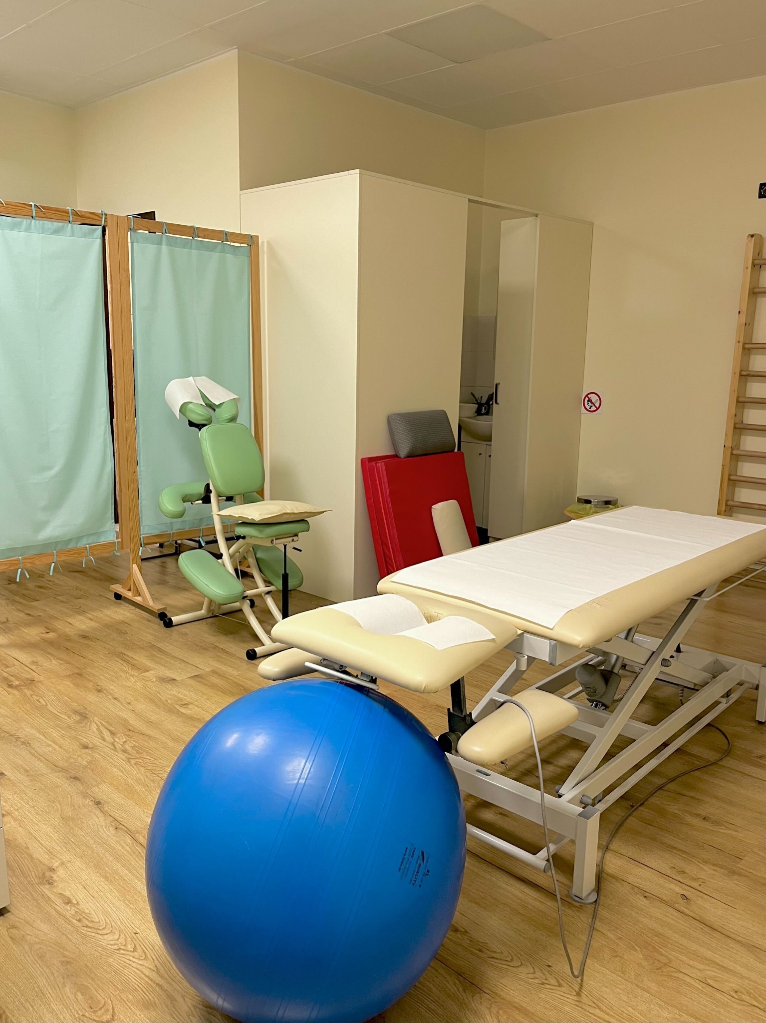 Jasne wnętrze gabinetu z profesjonalnym stołem do zabiegów, krzesłem do masażu, dużą niebieską piłką do ćwiczeń i drabinką gimnastyczną.
