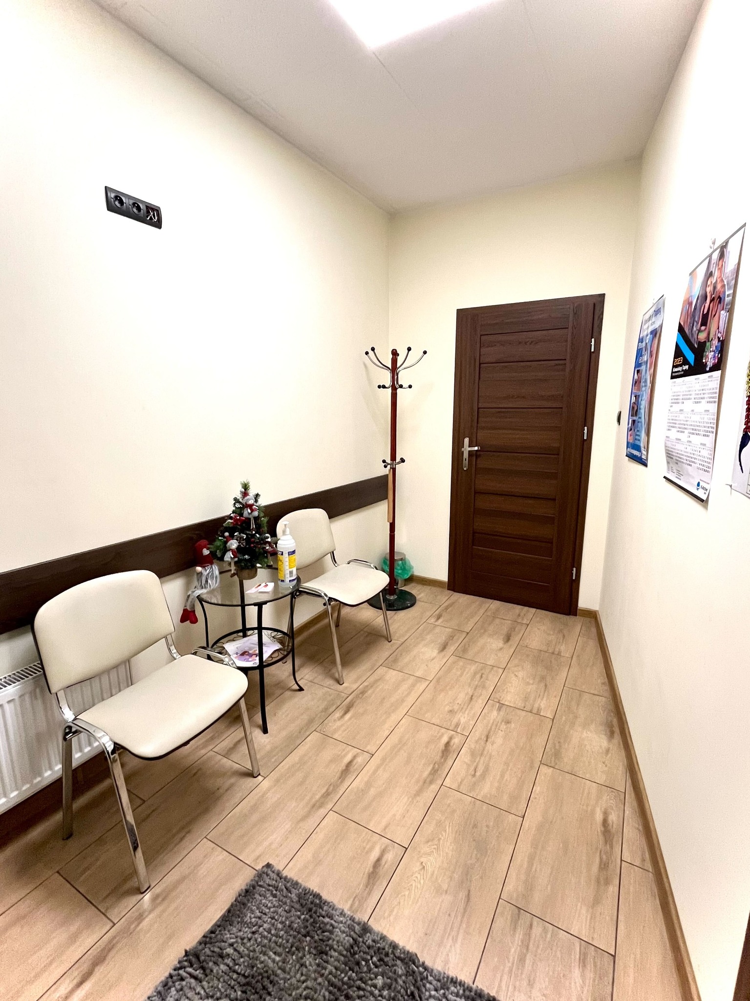 Jasne wnętrze gabinetu z kremowymi ścianami, drewnopodobną podłogą, dwoma krzesłami, małym stolikiem z dekoracją świąteczną, wieszakiem na ubrania i plakatami na ścianie.