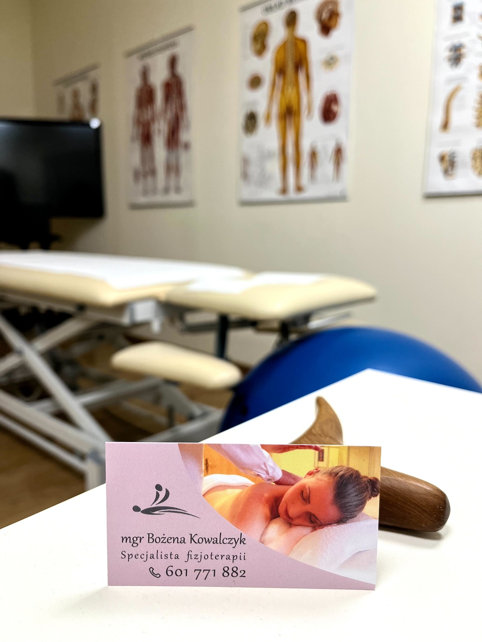 Wizytówka specjalisty z ujęciem pacjentki podczas zabiegu, drewniany masażer, stół do zabiegów i plakaty anatomiczne w tle.