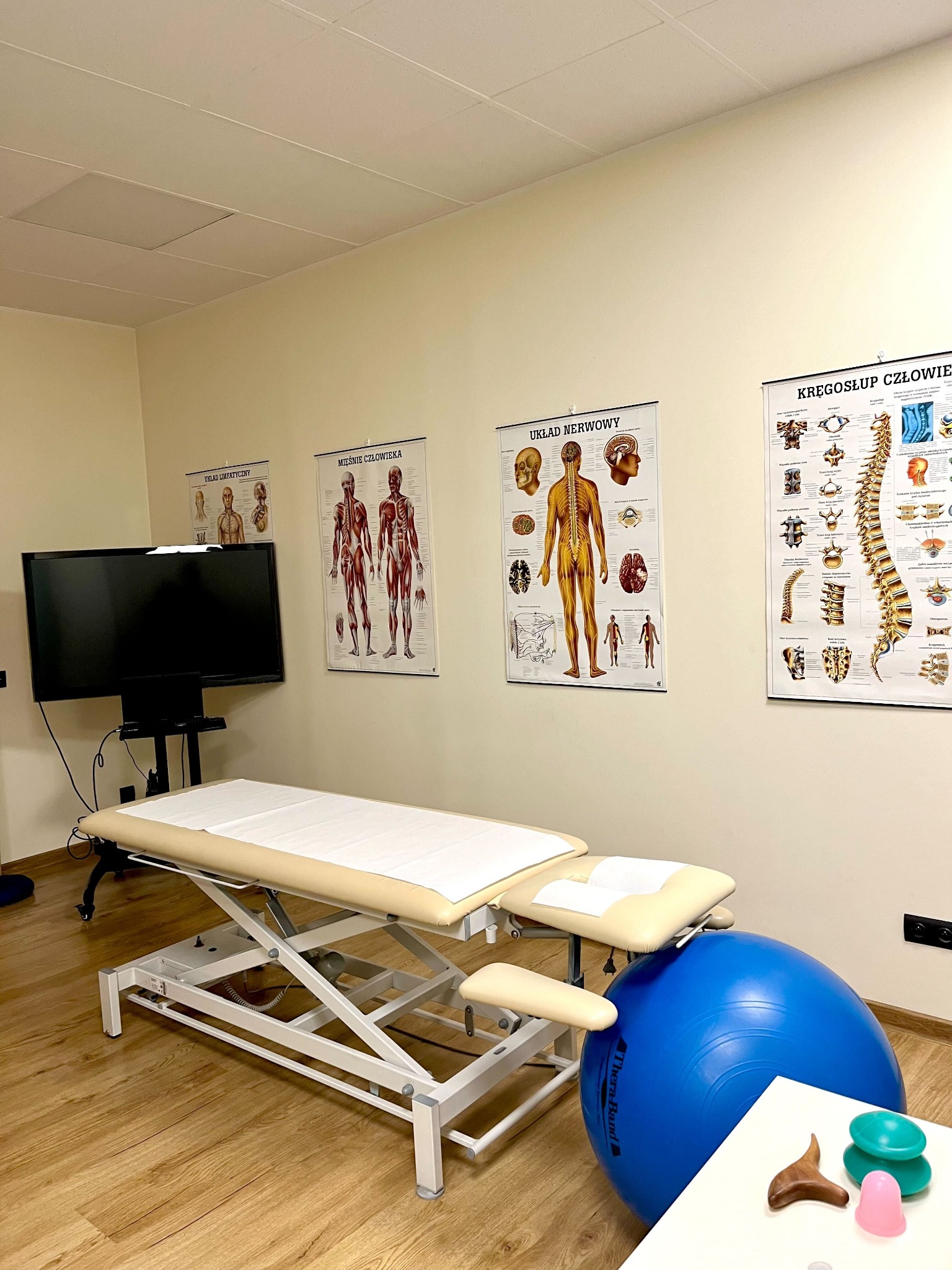 Gabinet z beżowym leżanką, niebieską piłką do ćwiczeń, bańkami chińskimi i plakatami anatomicznymi na ścianie.