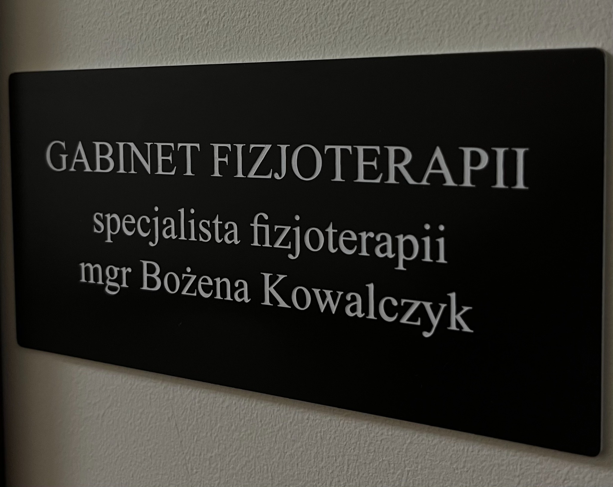 Tabliczka informacyjna na ścianie z napisem 'GABINET FIZJOTERAPII specjalista fizjoterapii mgr Bożena Kowalczyk' w kolorze białym na czarnym tle, lekko ukośne ujęcie.