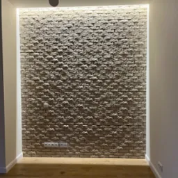 Ściana wykończona dekoracyjną cegłą z podświetleniem LED, widoczne gniazdka elektryczne i włącznik światła, drewniana podłoga