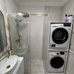 Jakub Solecki - Nowoczesna łazienka z kabiną prysznicową w złotej ramie, umywalką nablatową i pralko-suszarką Electrolux na tle marmurowych płytek. Minimalistyczny design.