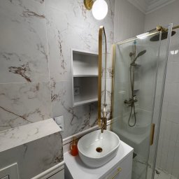 Jakub Solecki - Nowoczesna łazienka z marmurowymi płytkami, złotymi akcentami i owalną umywalką. Kabina prysznicowa ze złotymi okuciami. Ukryta szafka w ścianie.