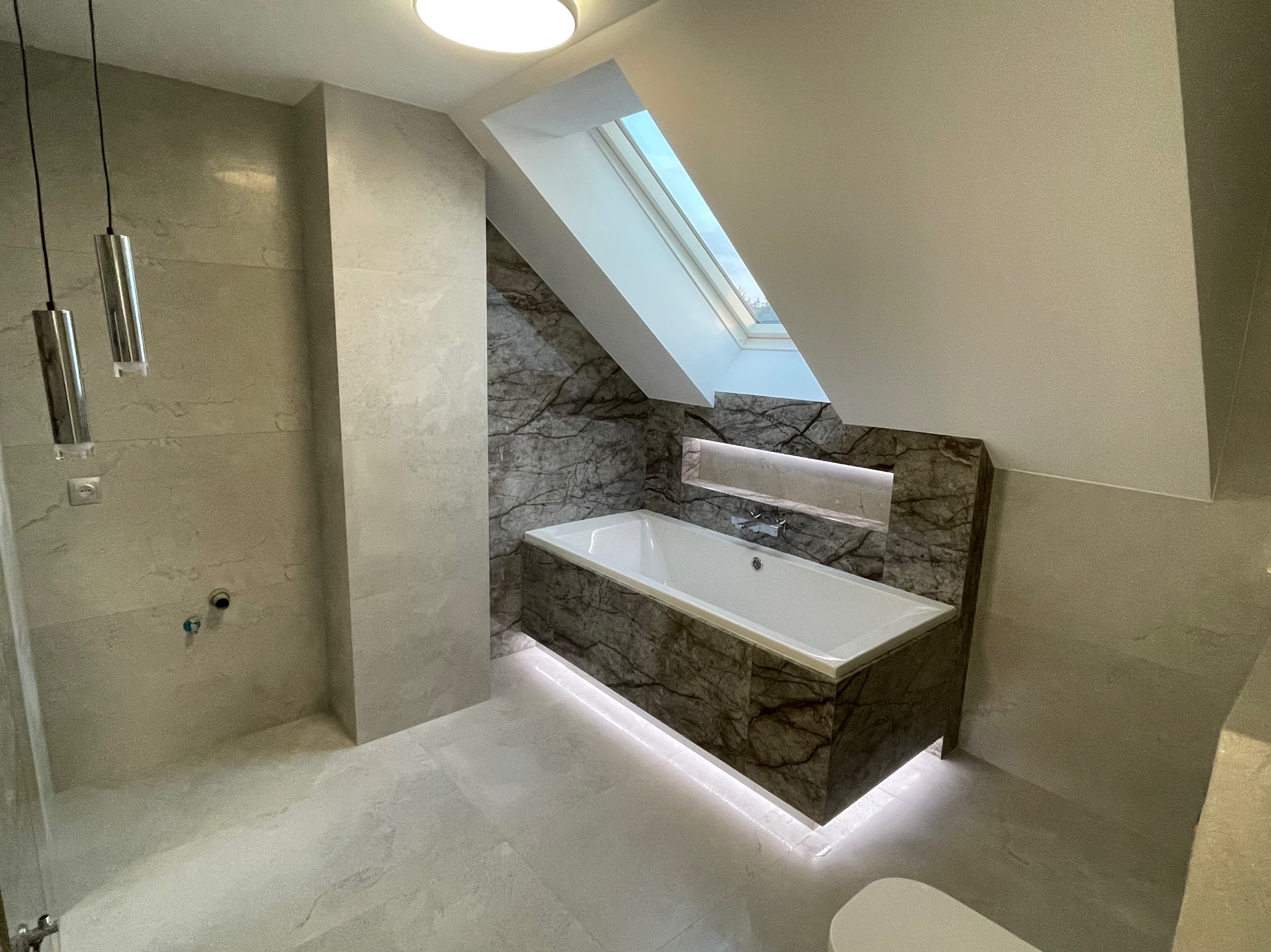 Nowoczesna łazienka na poddaszu z wanną obudowaną marmurem, podświetleniem LED od dołu i oknem dachowym, szare płytki na ścianach i podłodze, minimalistyczne lampy wiszące.