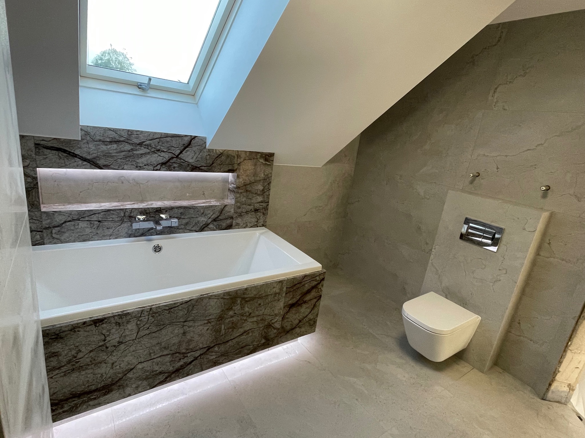 Nowoczesna łazienka na poddaszu z wanną obudowaną płytkami imitującymi marmur z podświetleniem LED, wiszącą toaletą i oknem dachowym.