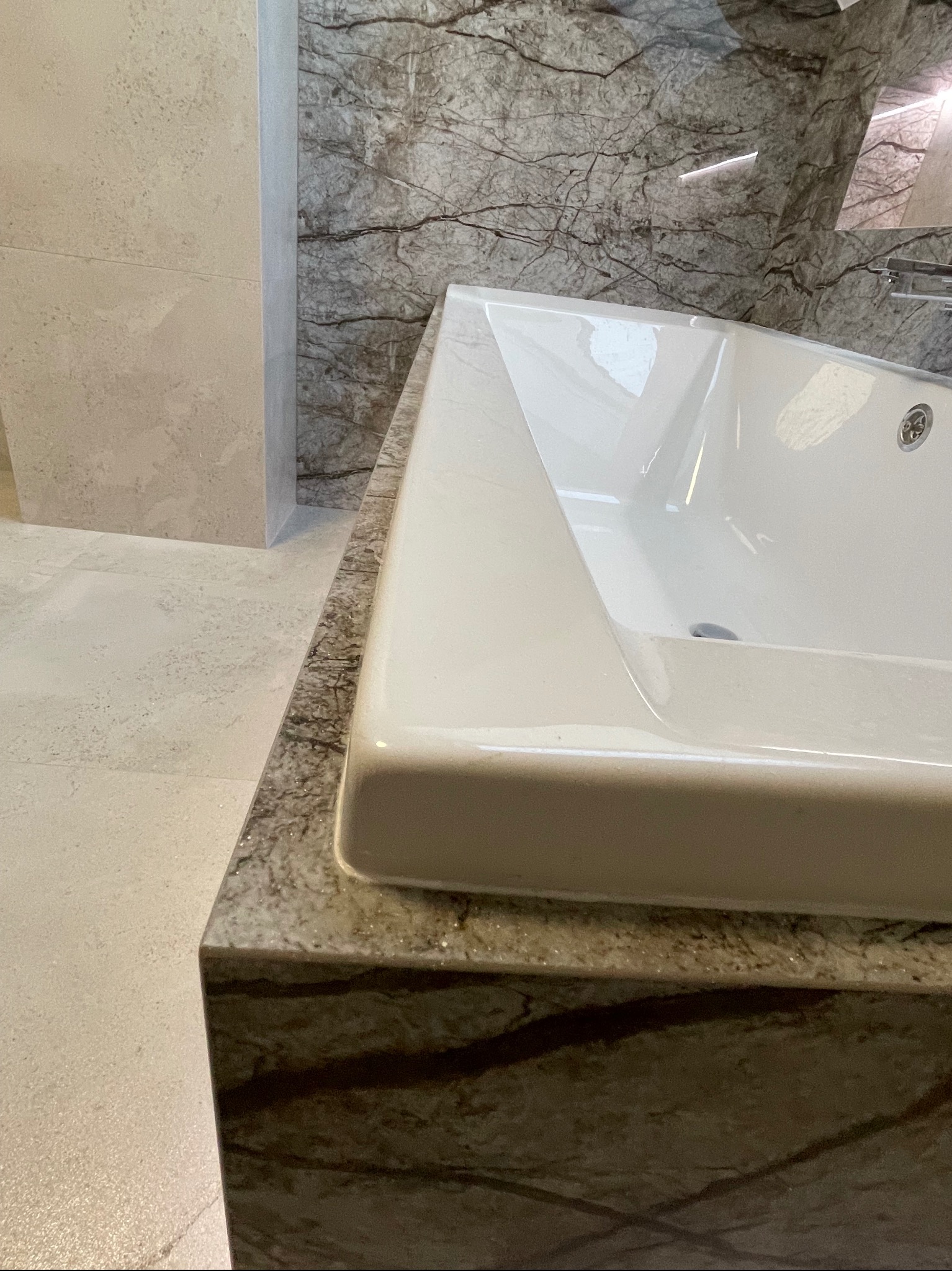 Biała umywalka nablatowa o geometrycznym kształcie na blacie z marmuru o wyraźnym, ciemnym żyłkowaniu, dopasowana do marmurowej ściany w tle.