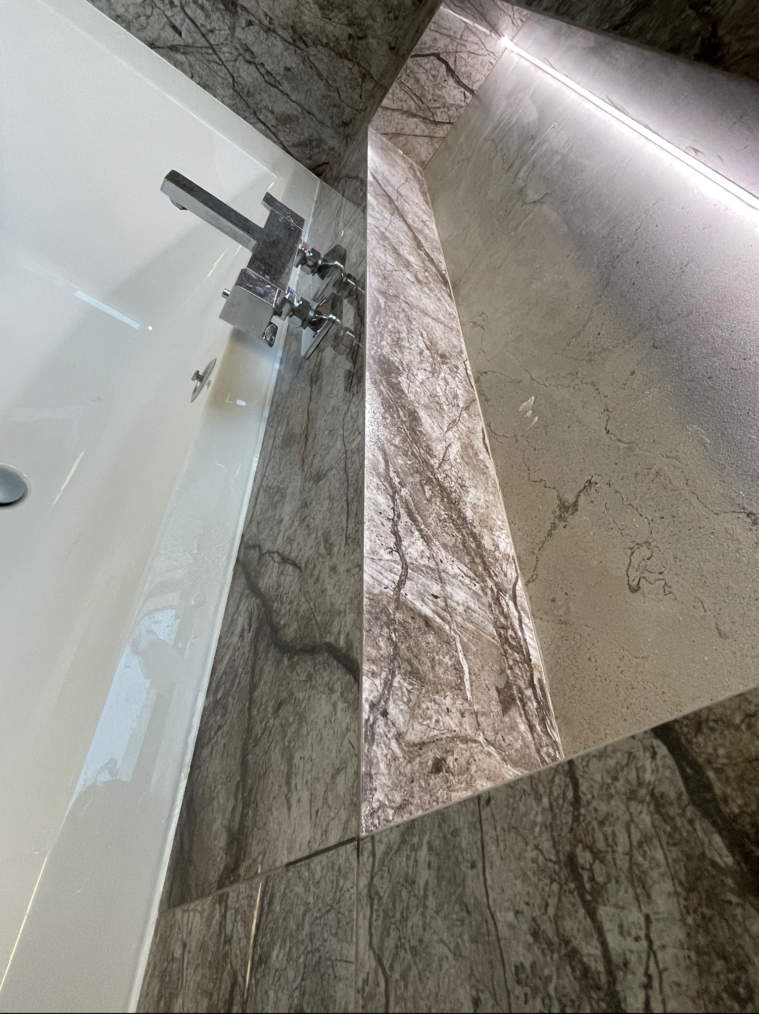 Biała wanna z chromowaną armaturą, obłożona płytkami imitującymi marmur w odcieniach szarości i brązu, z podświetlaną wnęką.