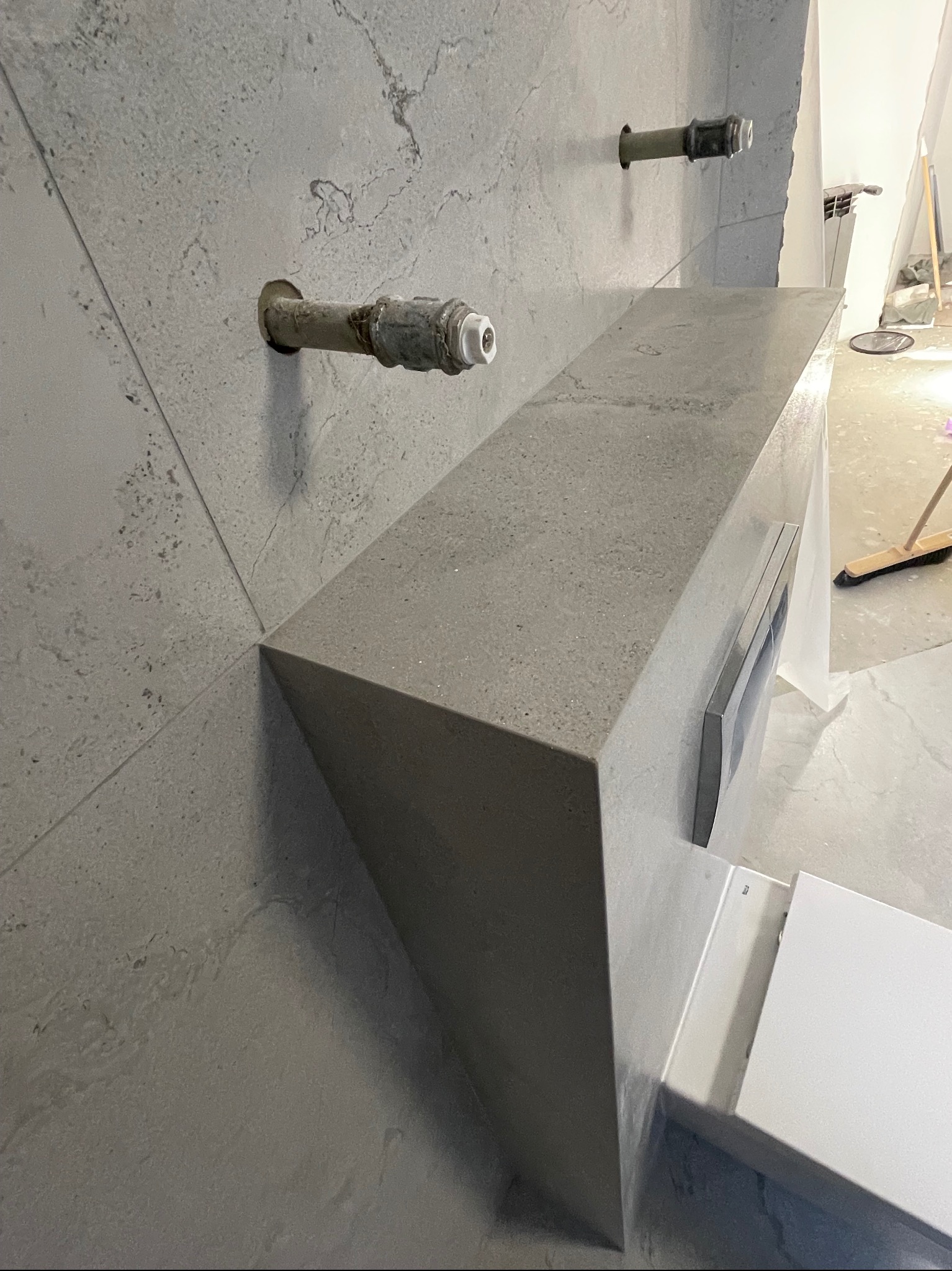 Szara, minimalistyczna umywalka z blatem z betonu architektonicznego, wystające rury instalacji wodnej w surowej łazience w trakcie remontu.