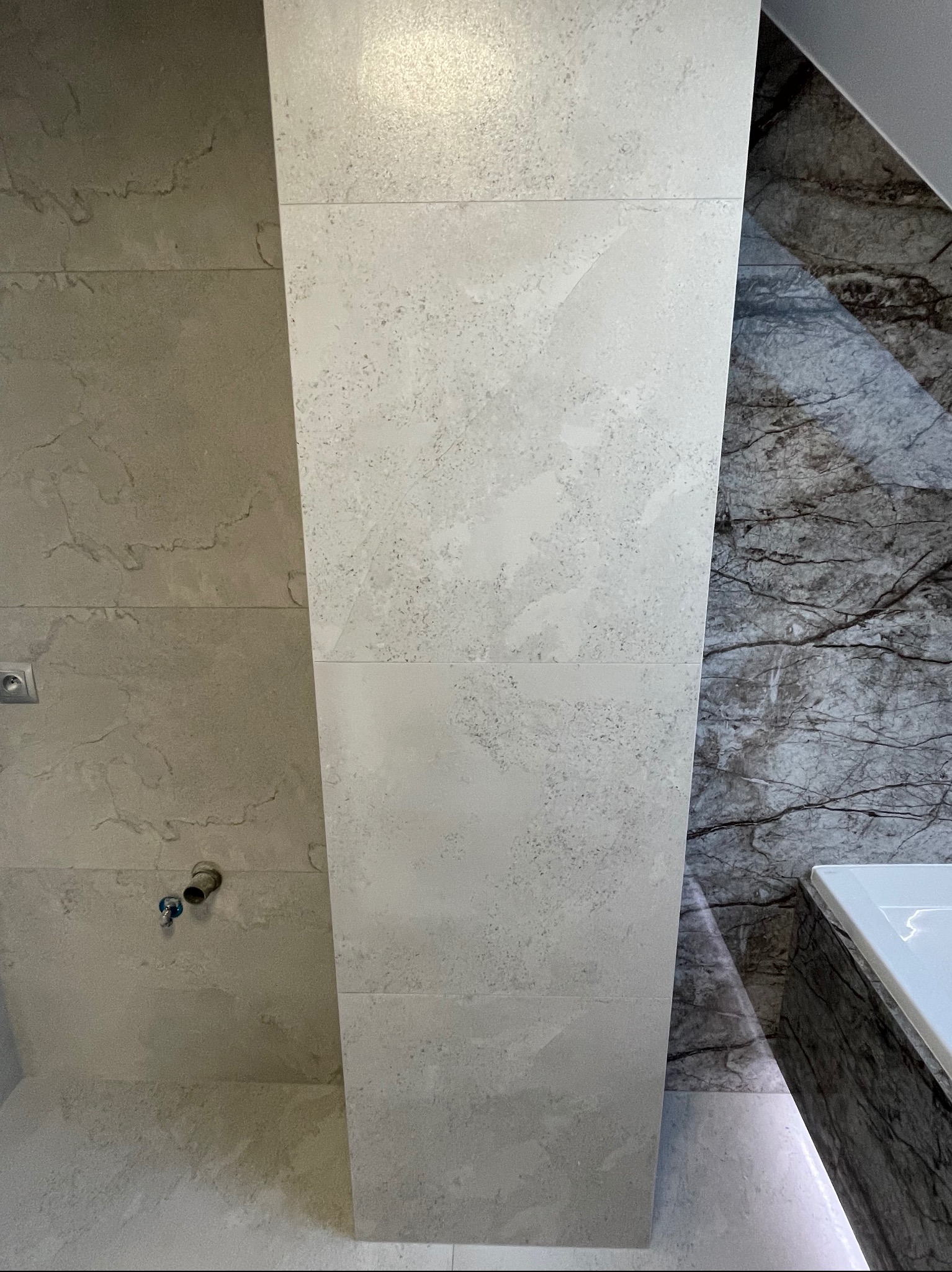 Fragment łazienki z wykończonymi ścianami: jasnoszare płytki imitujące kamień na jednej ścianie i ciemnoszara płyta marmurowa na drugiej, widoczny fragment instalacji wodnej.