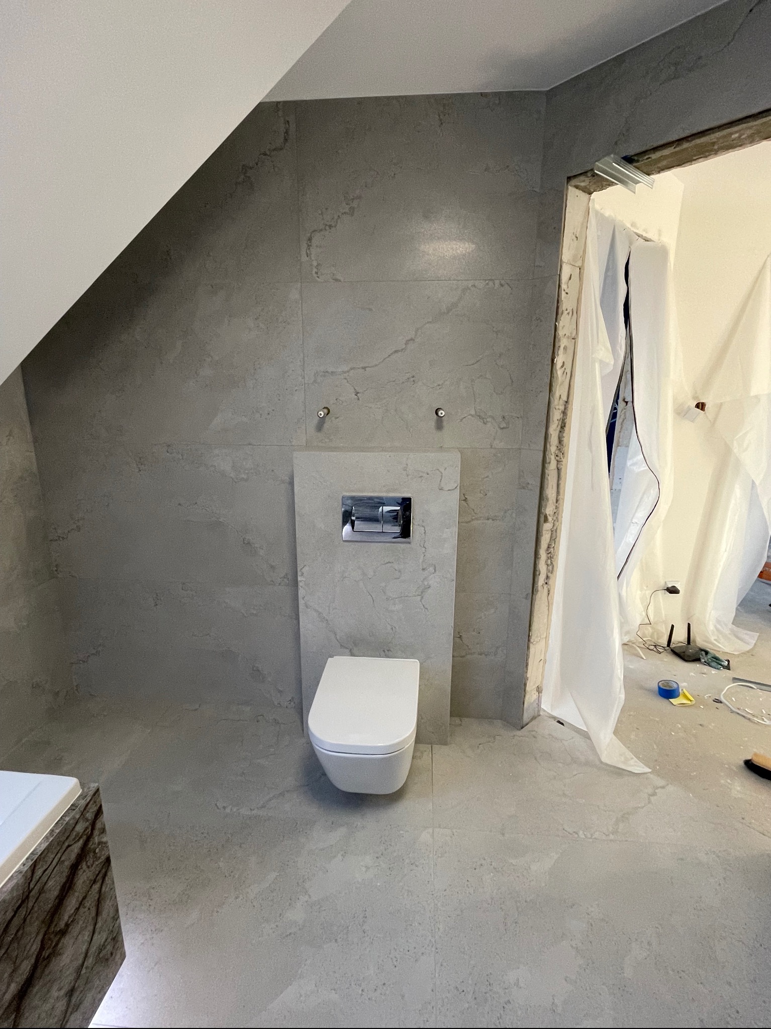Świeżo wykafelkowana łazienka na poddaszu z wiszącą toaletą, widoczne elementy instalacji i przygotowane wejście zabezpieczone folią malarską.