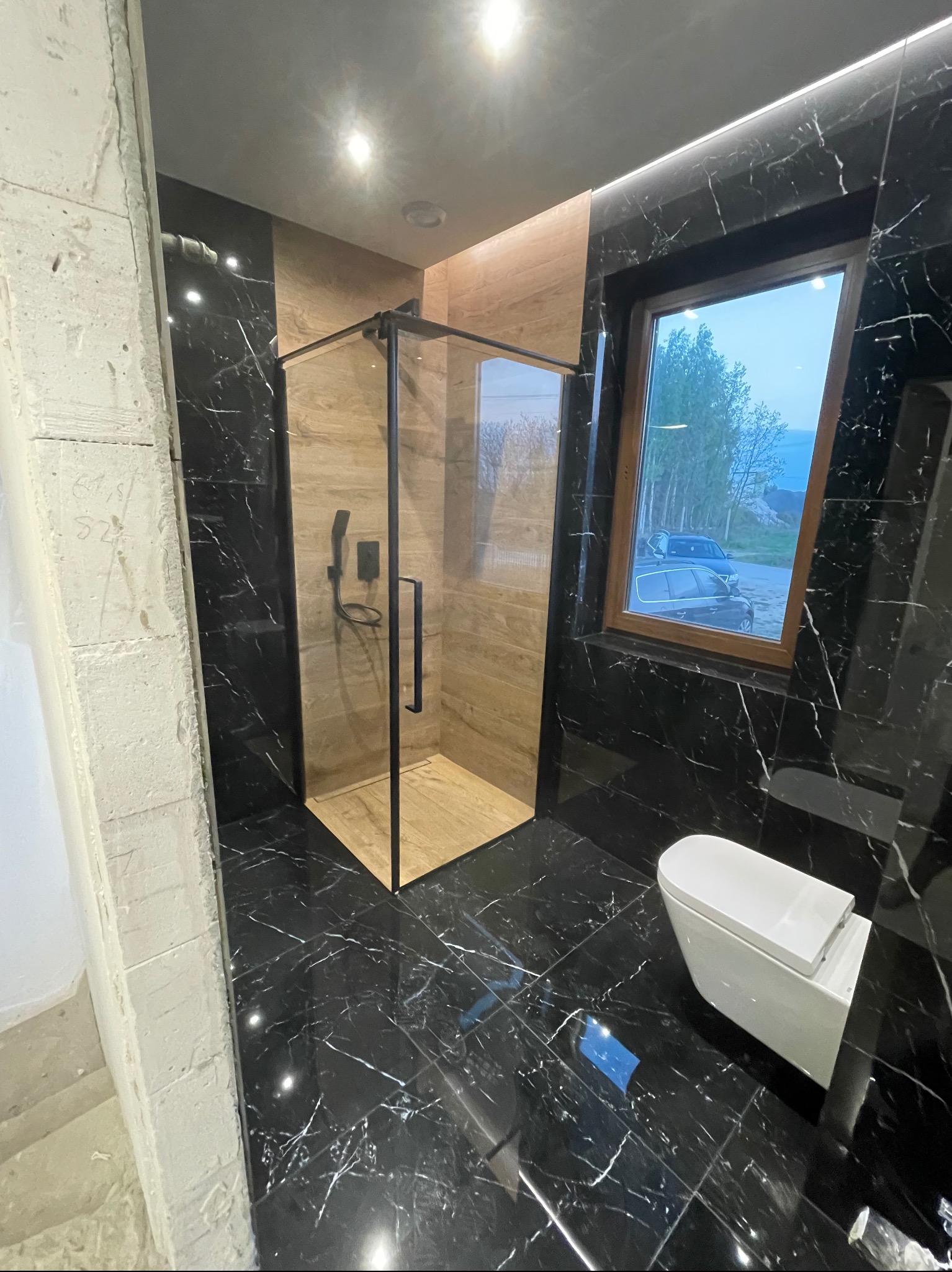 Nowoczesna łazienka z kabiną prysznicową typu walk-in, czarnymi marmurowymi płytkami z białymi żyłami, drewnopodobnymi panelami w kabinie, wiszącą toaletą i oknem z widokiem na zewnątrz.