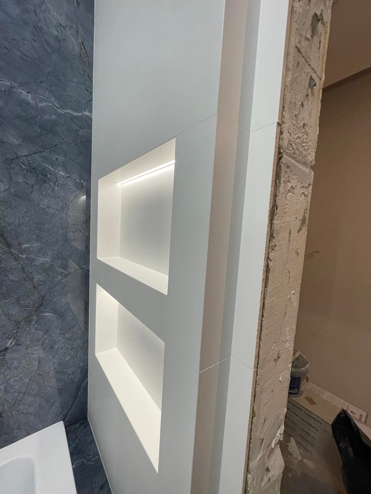 Wnęki z podświetleniem LED w białej zabudowie, kontrastujące z marmurową ścianą i surowym wykończeniem ściany obok, z widocznymi kartonami na podłodze.