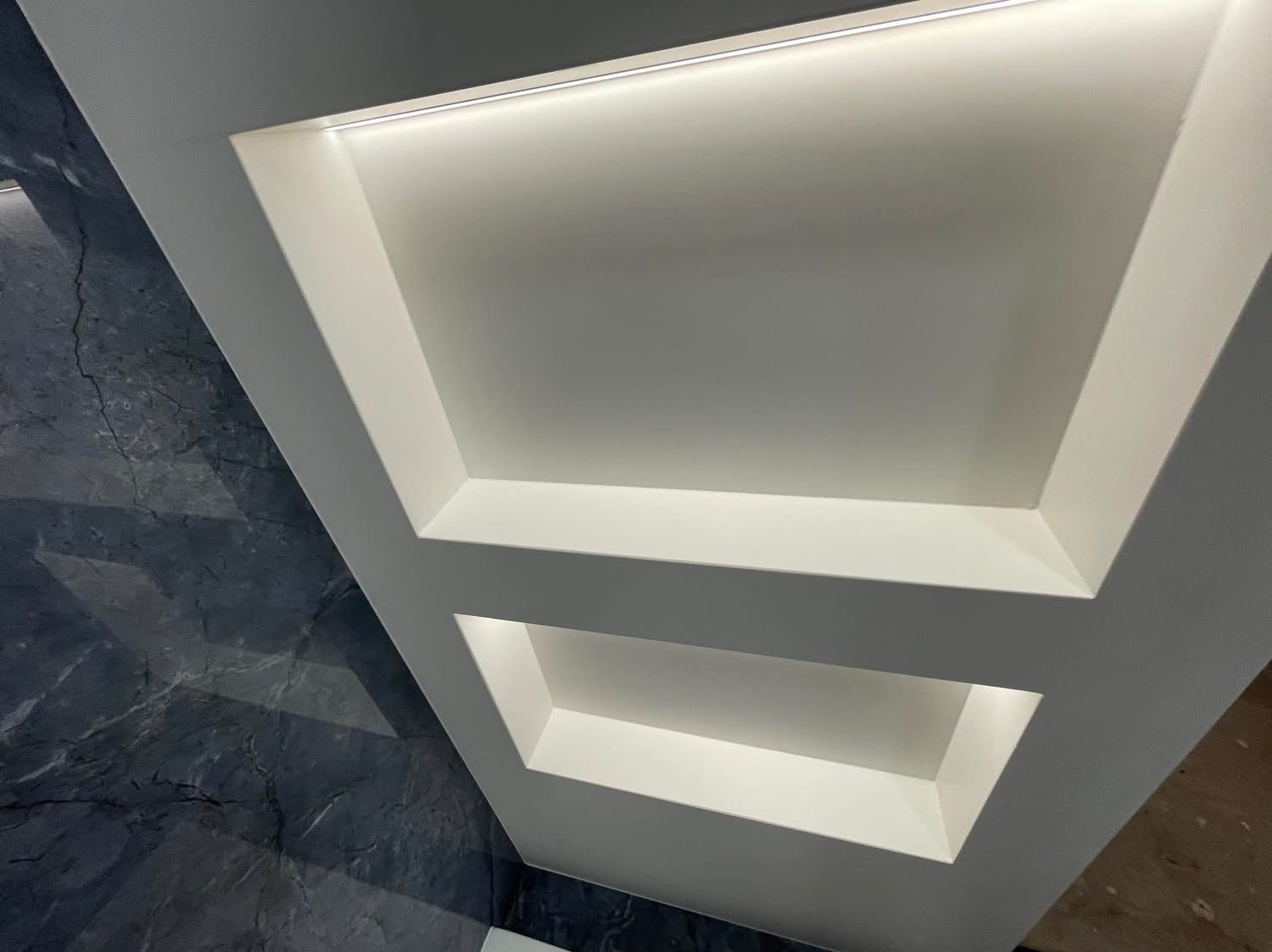 Nowoczesny sufit z wnękami oświetlonymi listwami LED, przylegający do ściany wykończonej płytami imitującymi ciemny marmur.