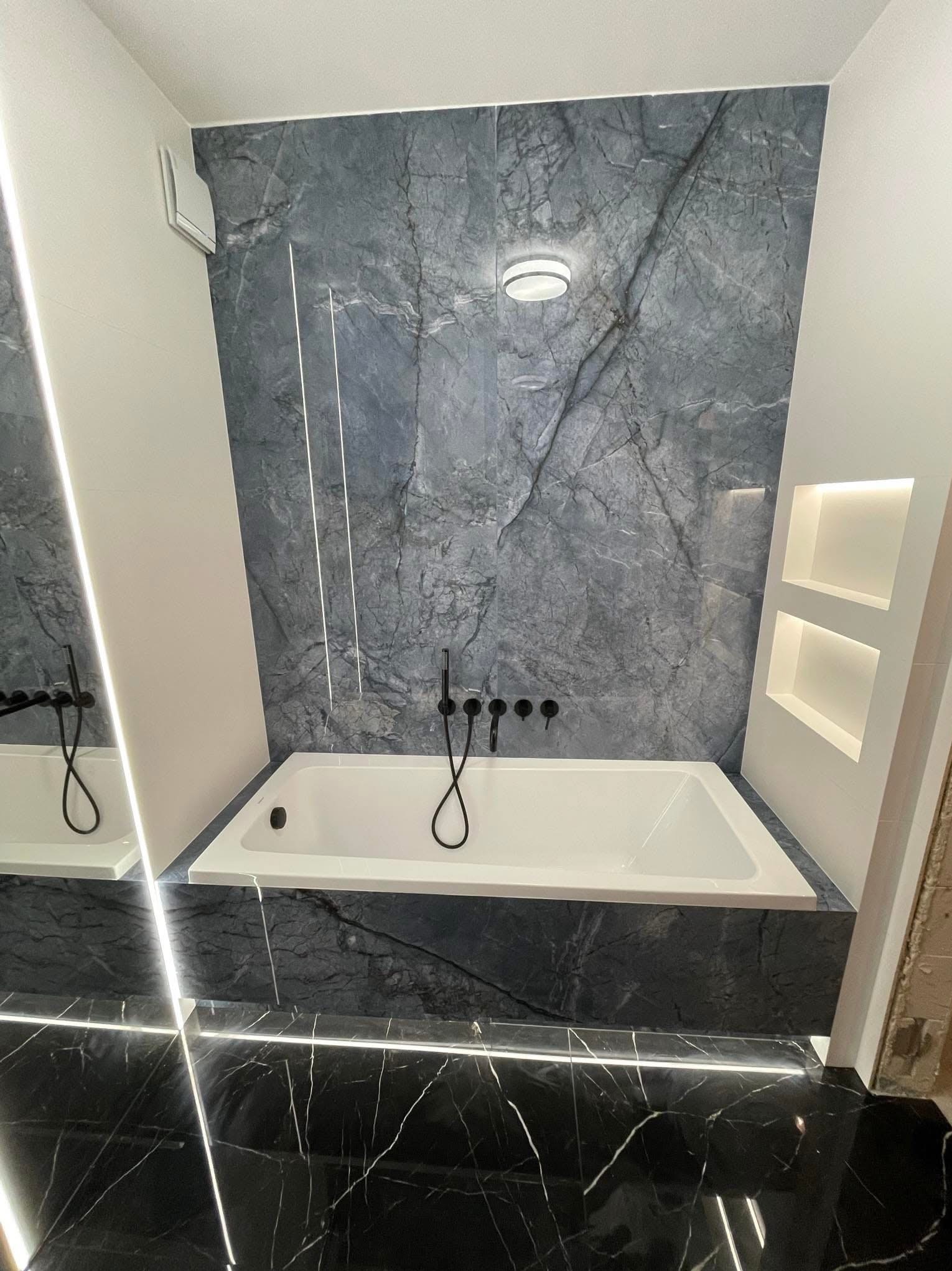 Nowoczesna łazienka z wanną obłożoną czarnym marmurem z białym żyłowaniem, ściana z szarego marmuru z oświetleniem LED, czarna armatura i podświetlane półki w ścianie.